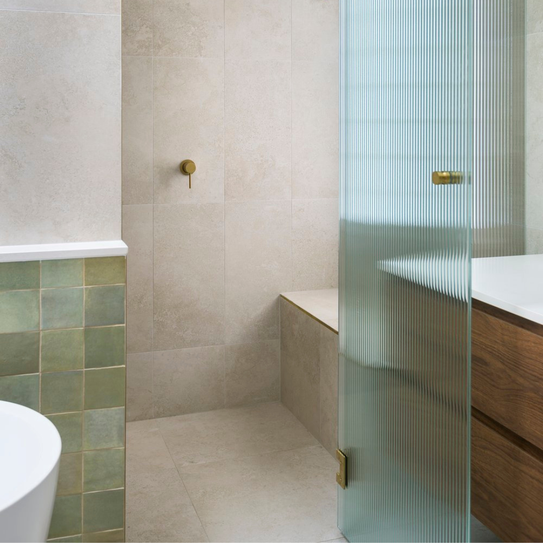 Modern bathroom with beige tiled walls and floor, partial green tiled wall, glass shower door with brass hardware, and wooden vanity with a white countertop.