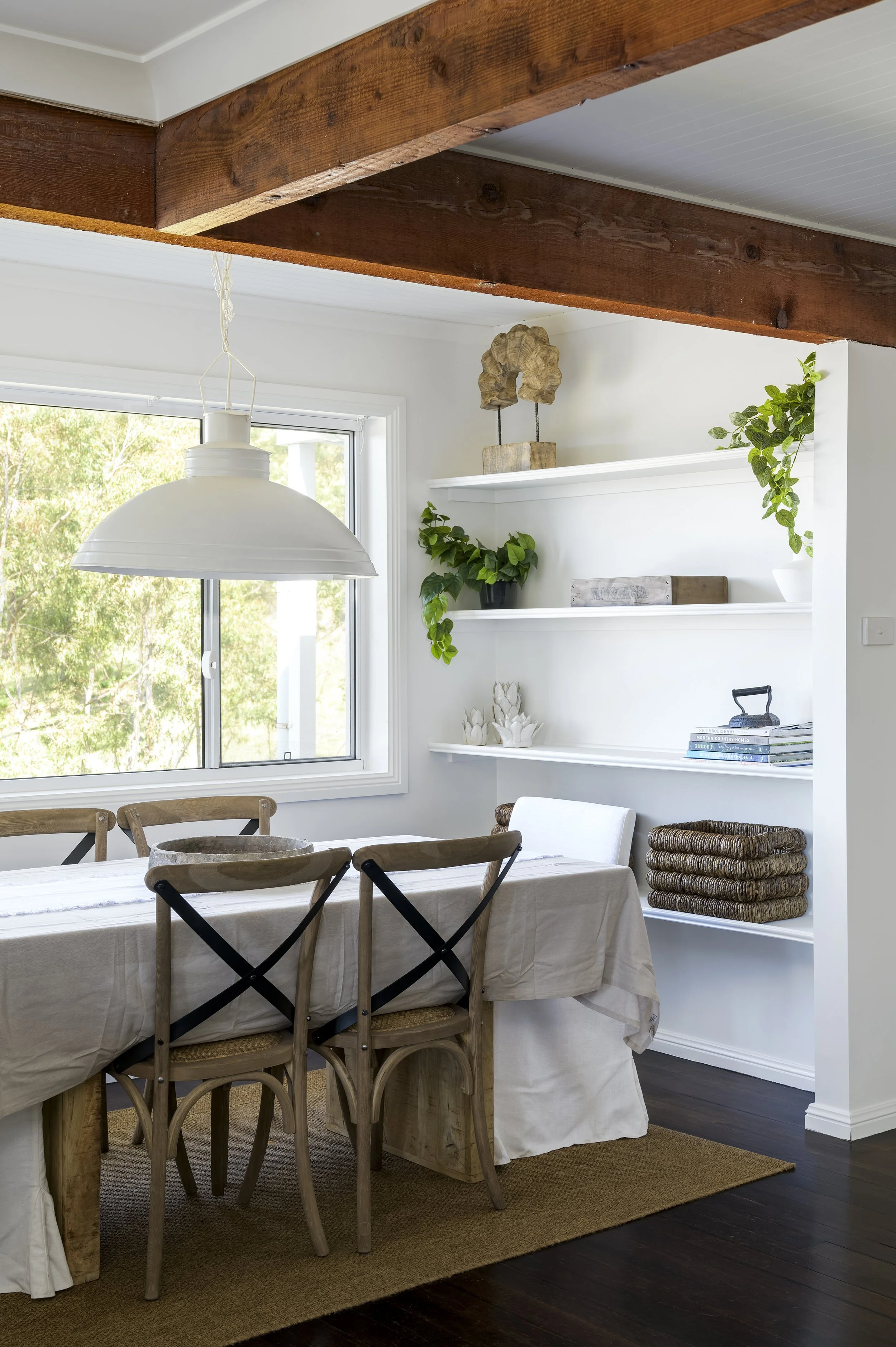 A dining room with a wooden table covered with a white tablecloth, surrounded by wooden chairs, a window with lush green trees outside, white shelves decorated with plants and art, and a large white pendant light overhead.
