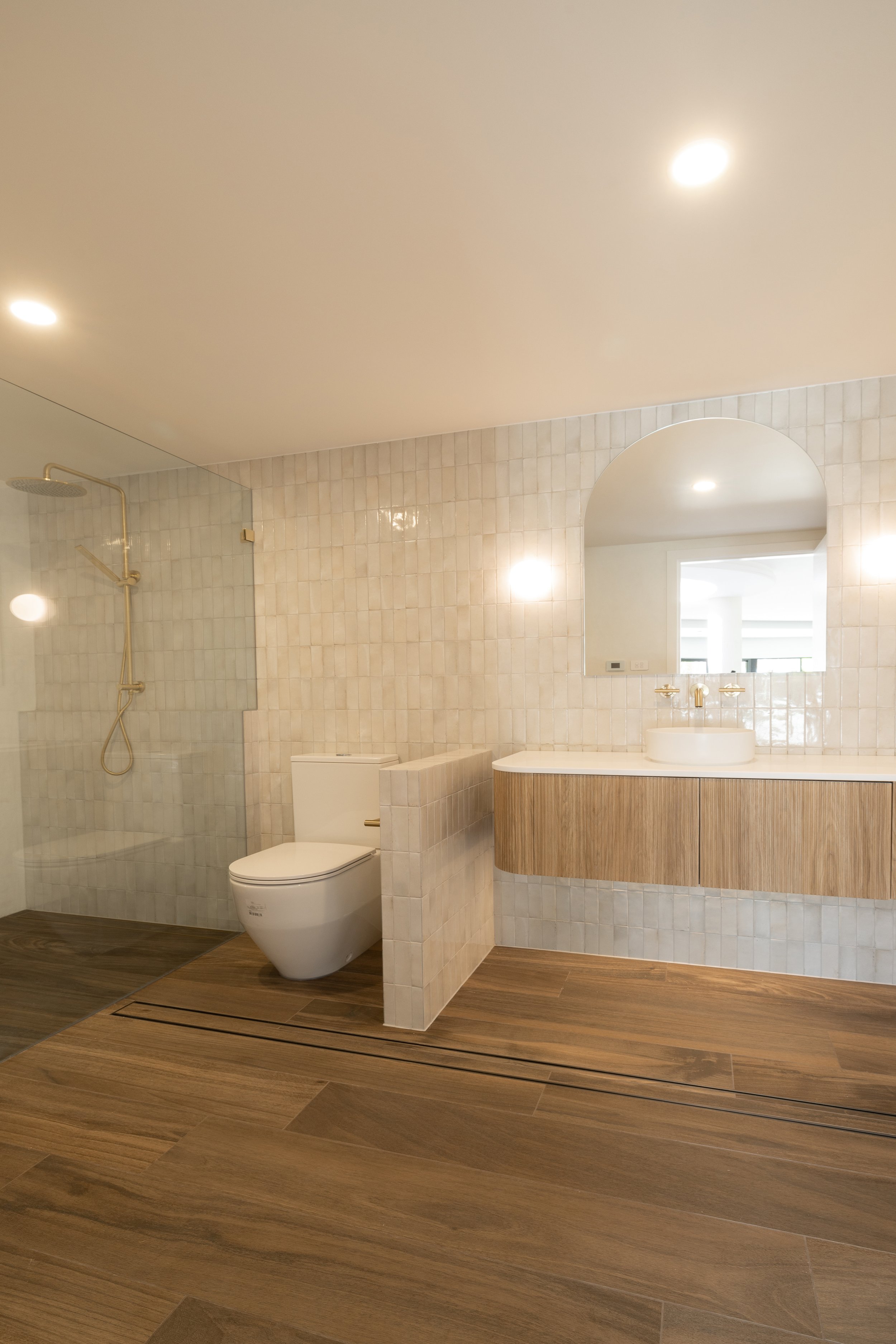 Modern bathroom with wooden flooring, a walk-in shower with glass enclosure, a toilet, a floating vanity with a round vessel sink, and a large mirror