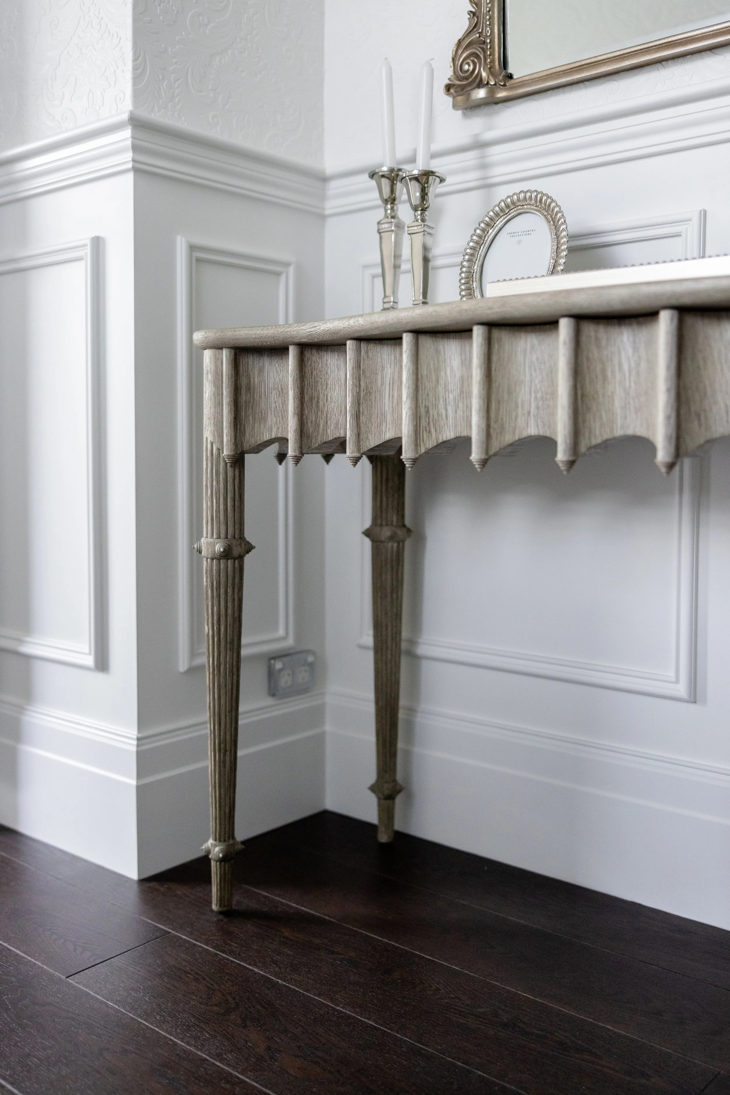 Decorative console table with candlesticks and picture frames in a white room with dark wood flooring.