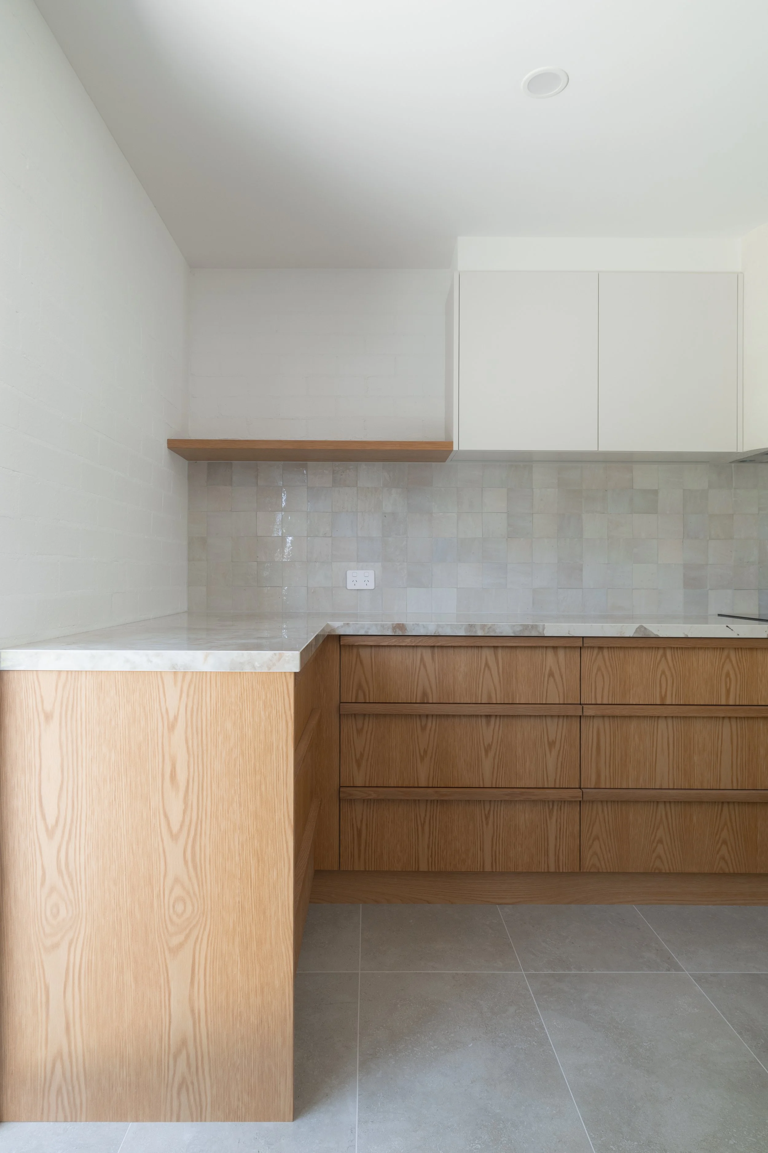 Minimalist kitchen with wood cabinetry, a marble countertop, beige tiled backsplash, white upper cabinets, and a small wooden shelf.