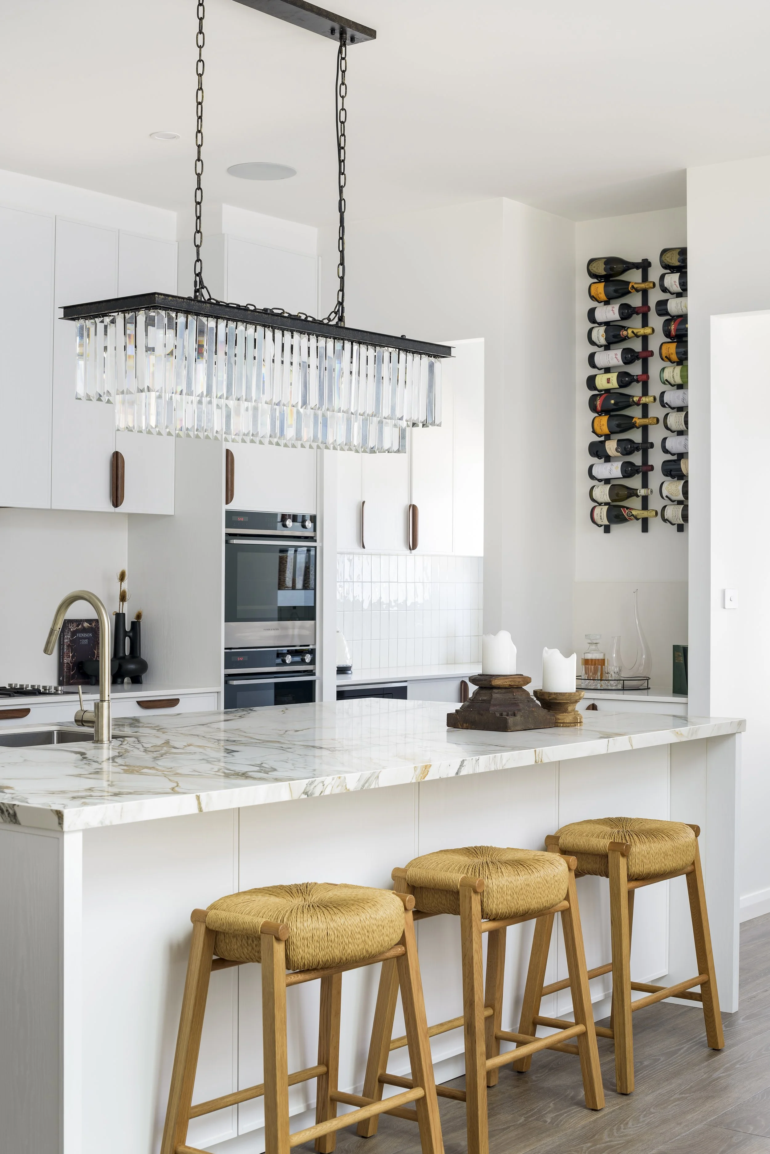 Modern kitchen with white cabinets, marble island, wine rack on the wall, and three wooden bar stools with woven seats.