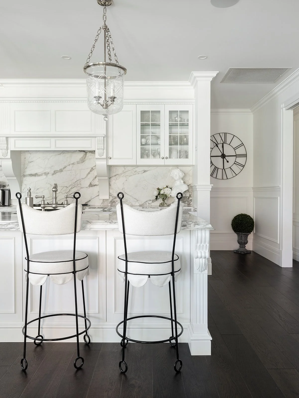 A white kitchen with marble countertops and backsplash, including a chandelier above the island, black-framed white barstools, a decorative clock on the wall, and dark hardwood floors.