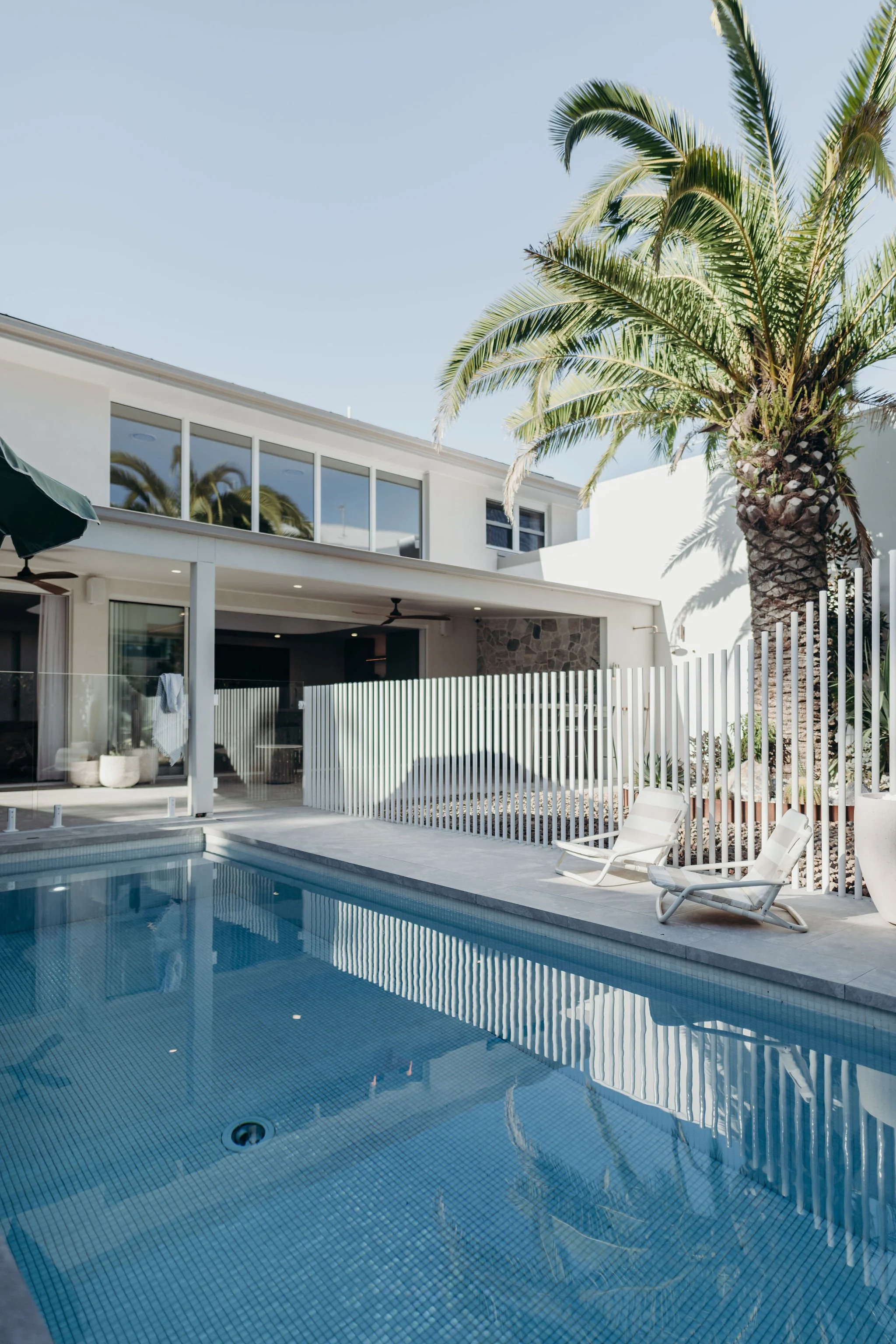 A modern backyard with a swimming pool, two white lounge chairs, a white fence, and a tall palm tree in front of a white house with large windows.