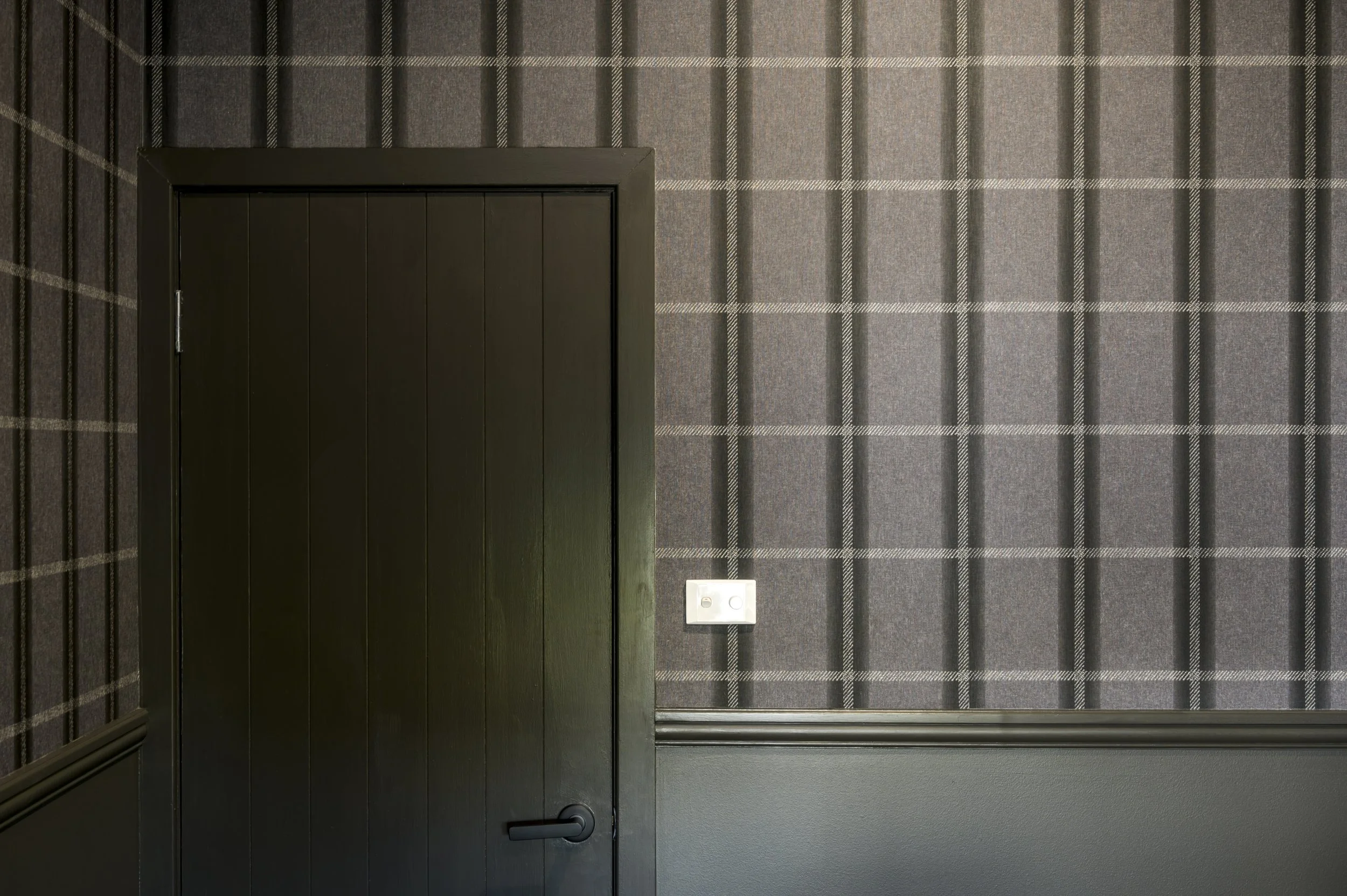 A black door with a handle on a gray wall with plaid-patterned wallpaper, and an electrical outlet on the wall to the right of the door.