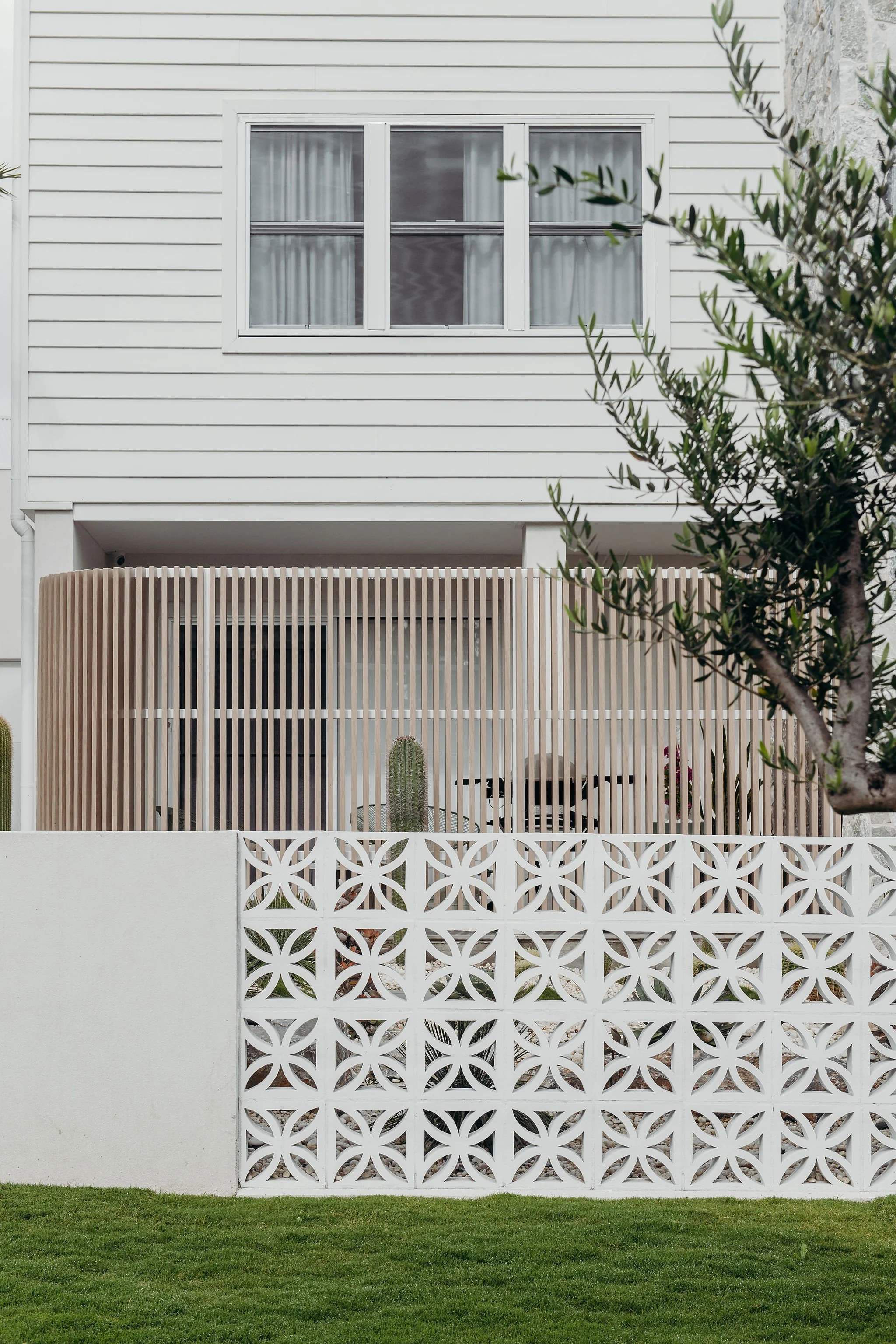 White house with a window, a decorative wooden fence on a porch, and a white patterned wall in front, with a cactus and a tree in the yard.