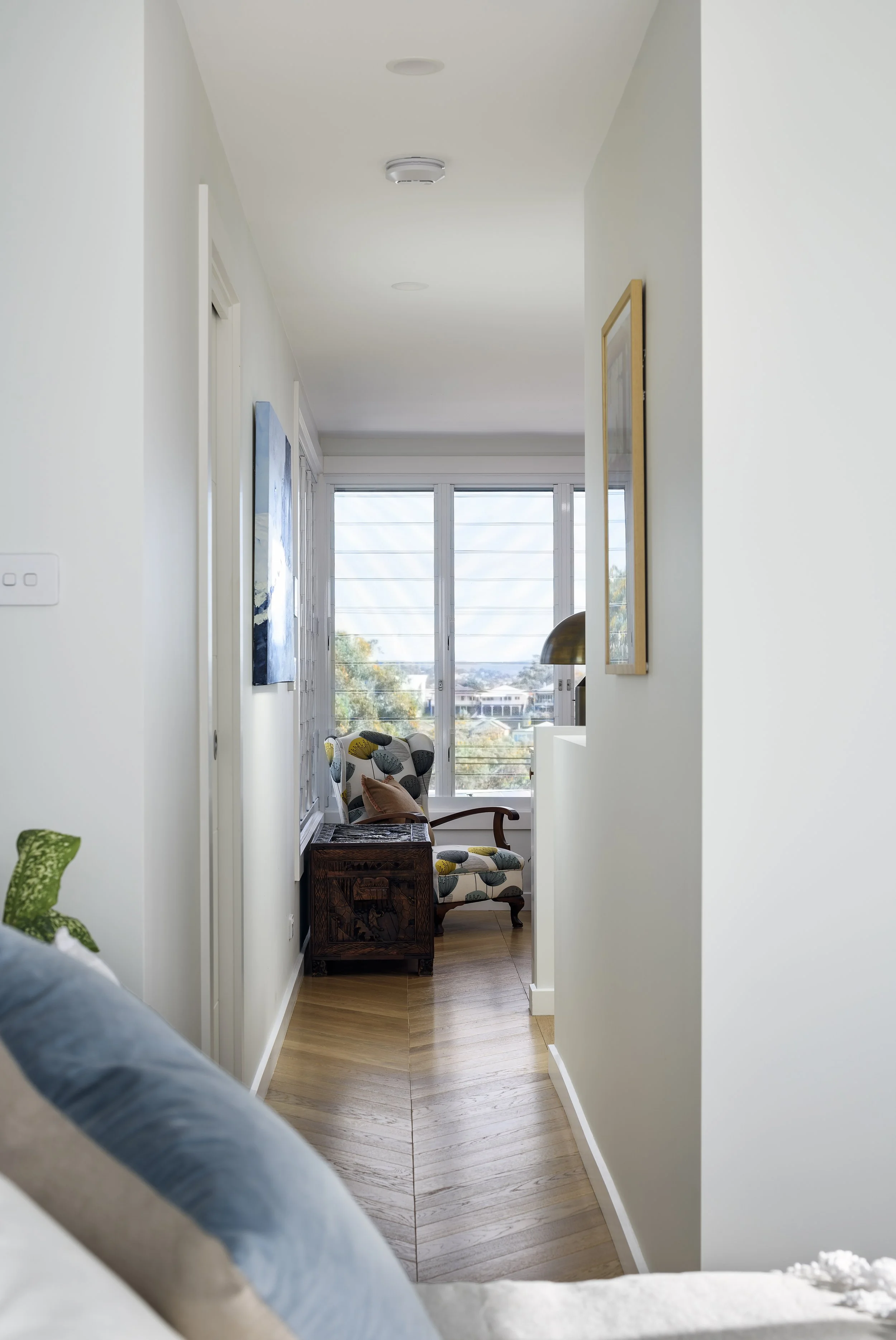 A narrow hallway leading to a sitting area with a window view of trees and buildings outside. The sitting area has a patterned armchair, a small wooden cabinet, and a metallic table lamp. The hallway has white walls and wooden flooring.