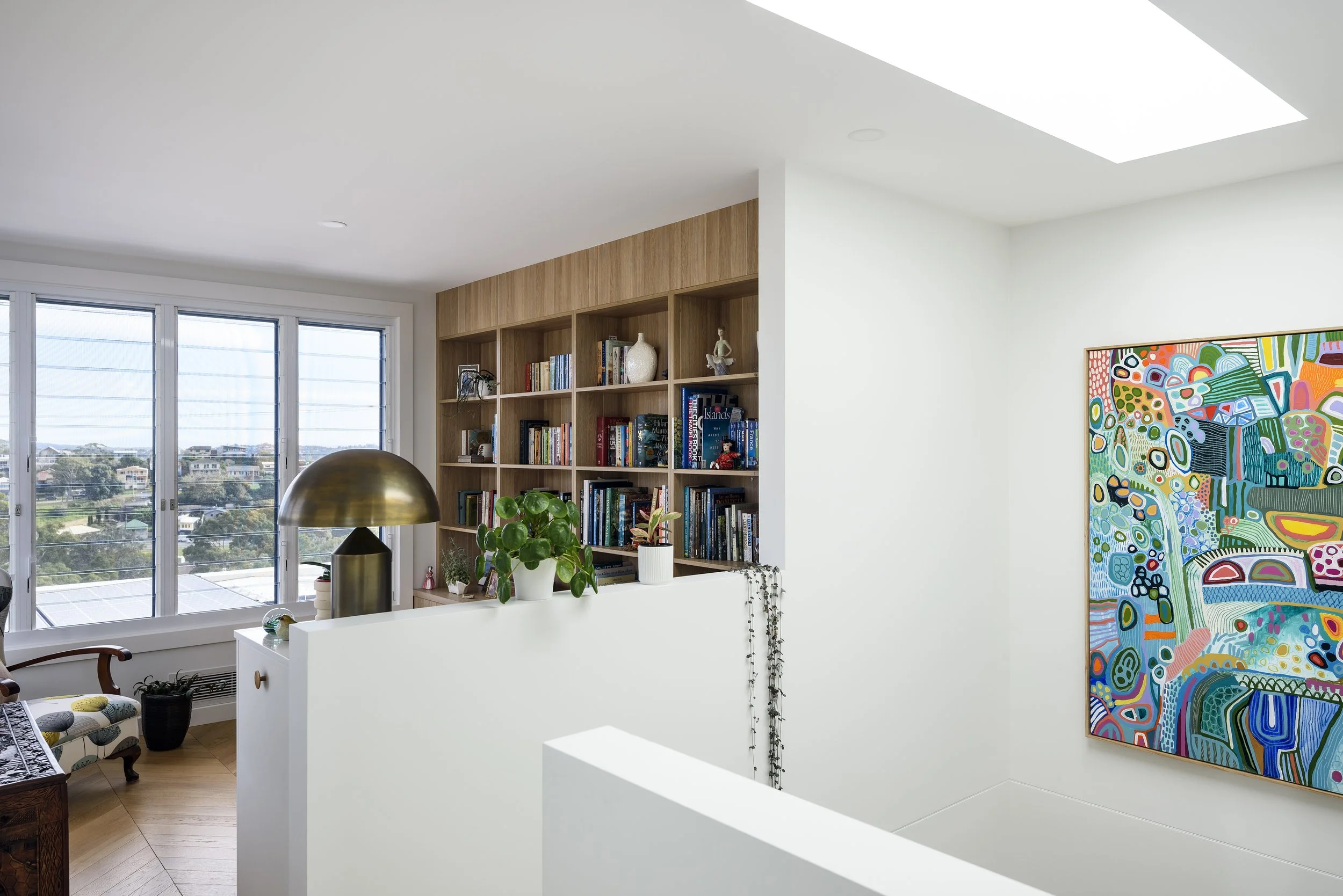 Bright living room with large windows, wooden bookshelf filled with books and decorative objects, colorful abstract artwork on a white wall, and a brass table lamp on a white counter with potted plants.