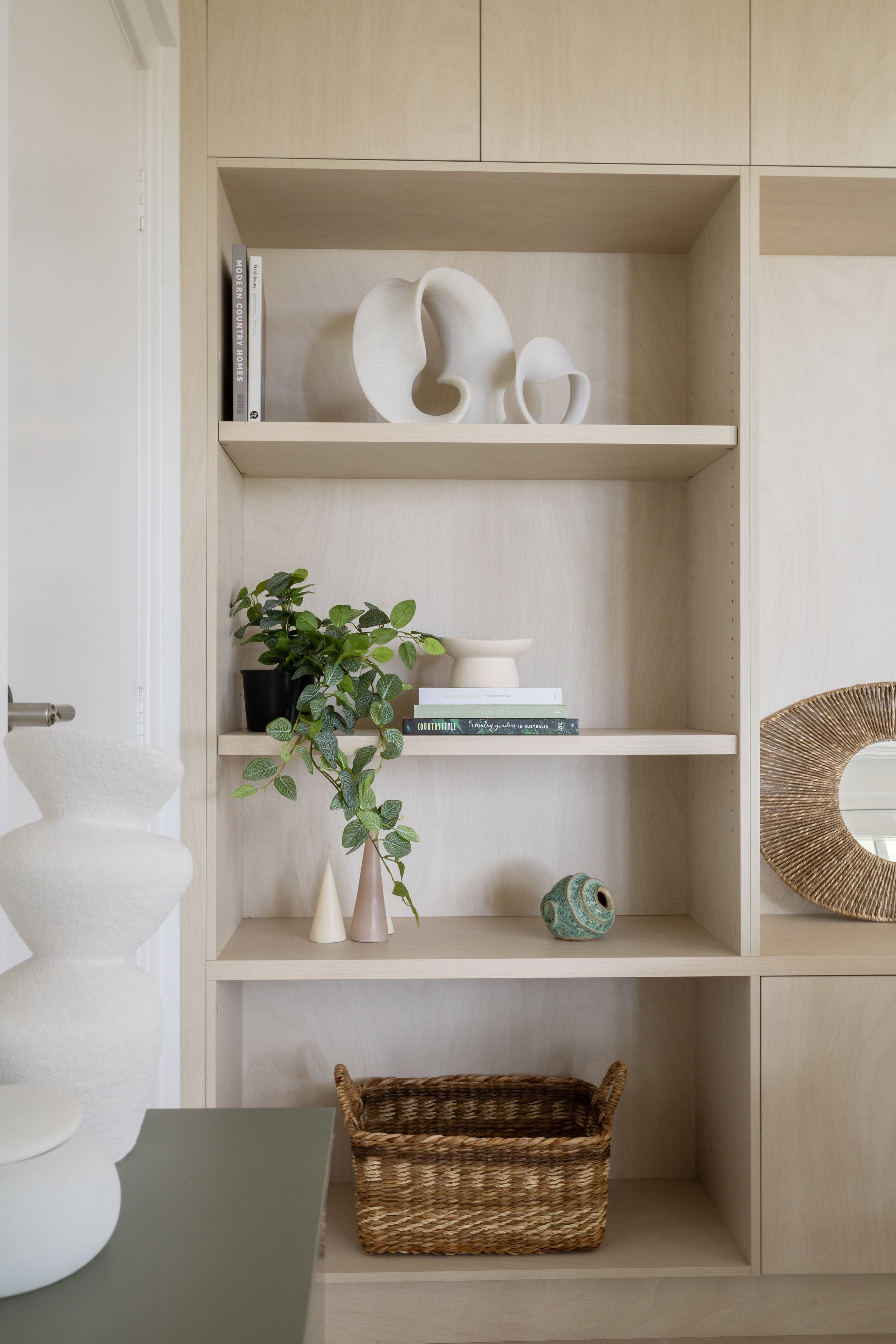 A wooden bookshelf with decorative items, including abstract white sculptures, green plants, books, small vases, and a woven basket inside.