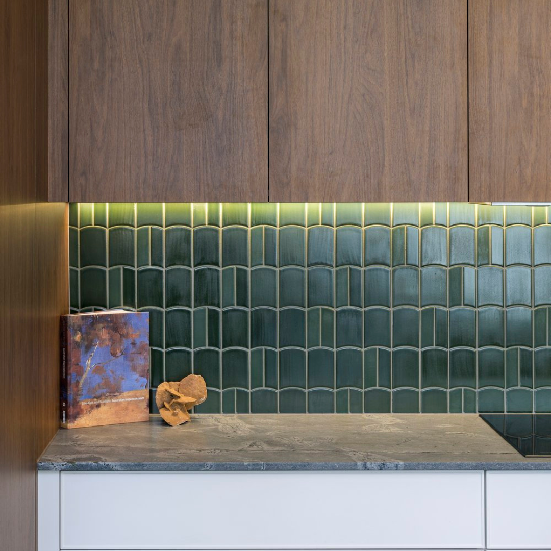 Kitchen countertop with green tiled backsplash, wooden cabinets above, and a book with driftwood decoration on the left side.