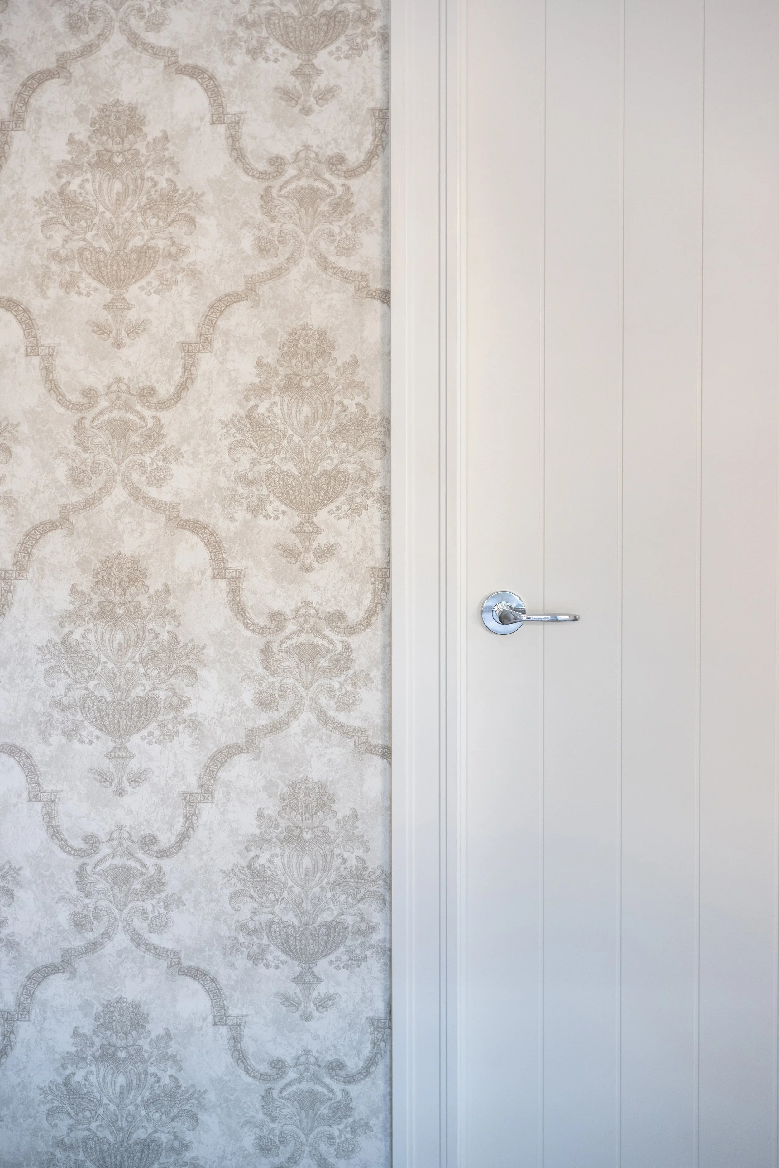 A white door with a modern lever handle next to a wall with beige floral-patterned wallpaper.