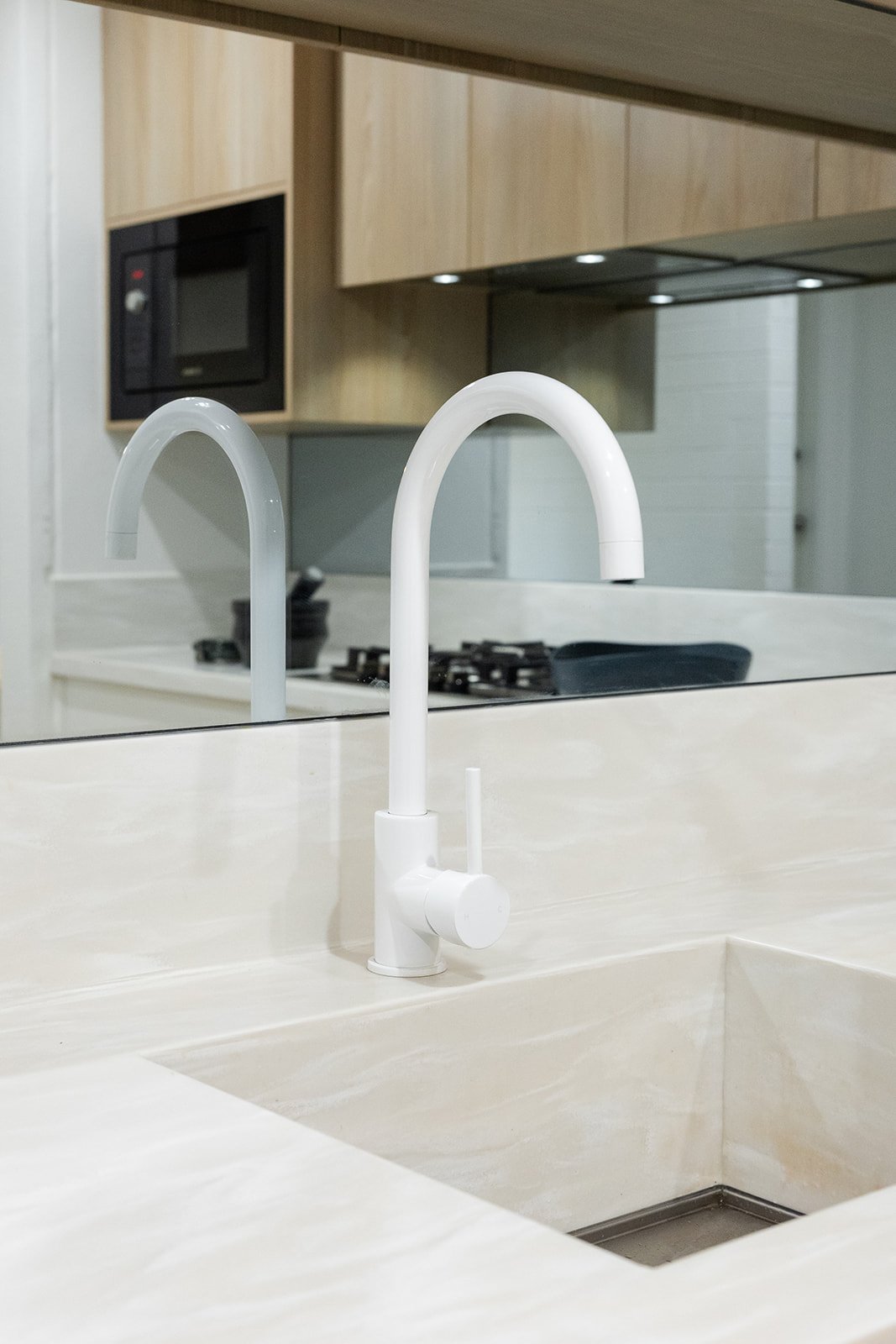 Close-up of a modern white kitchen sink faucet with a curved spout, reflected in a mirror, with a stovetop, microwave, and wooden cabinets in the background.