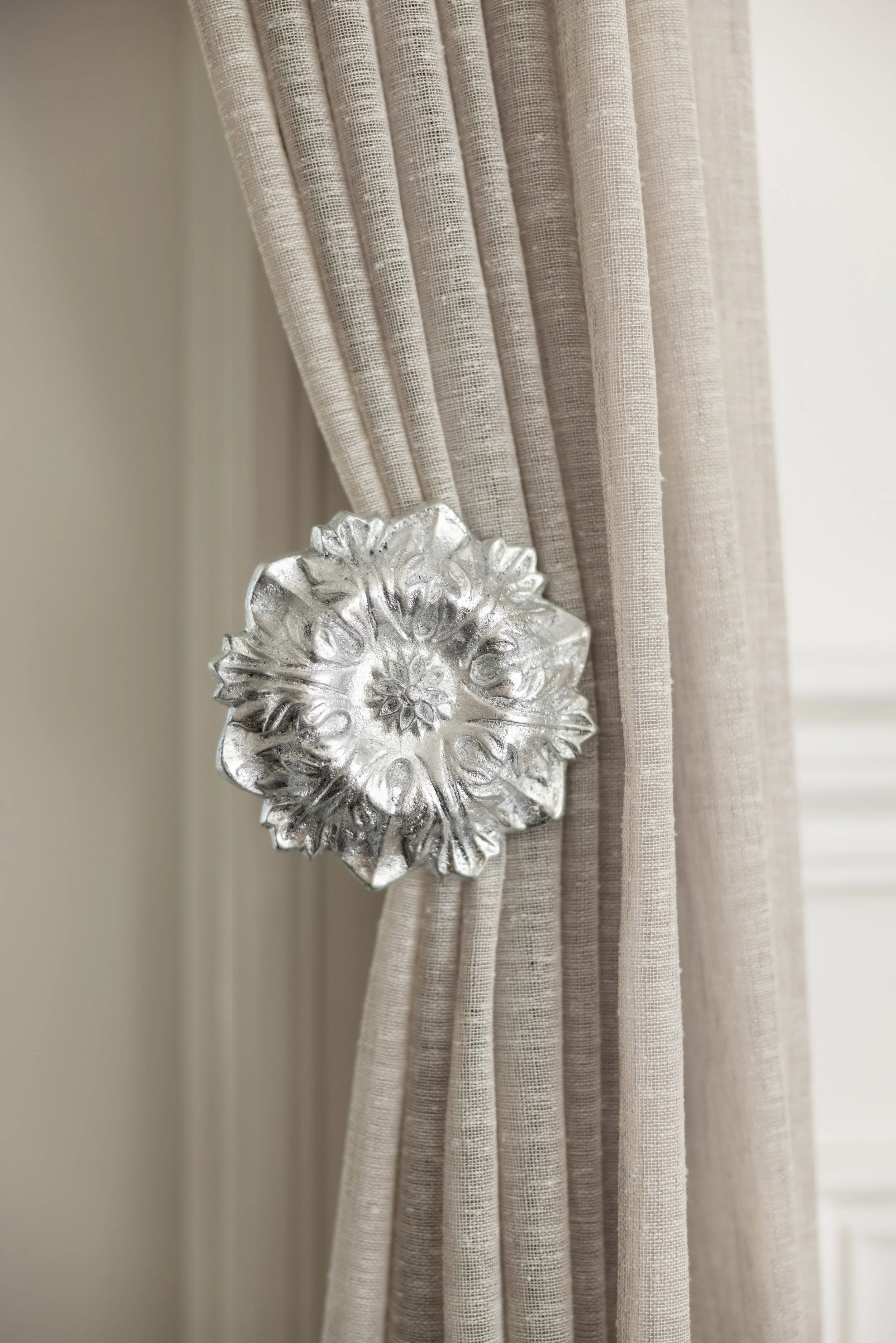 Beige curtains held back with a large, decorative, metallic silver tieback with floral patterns.