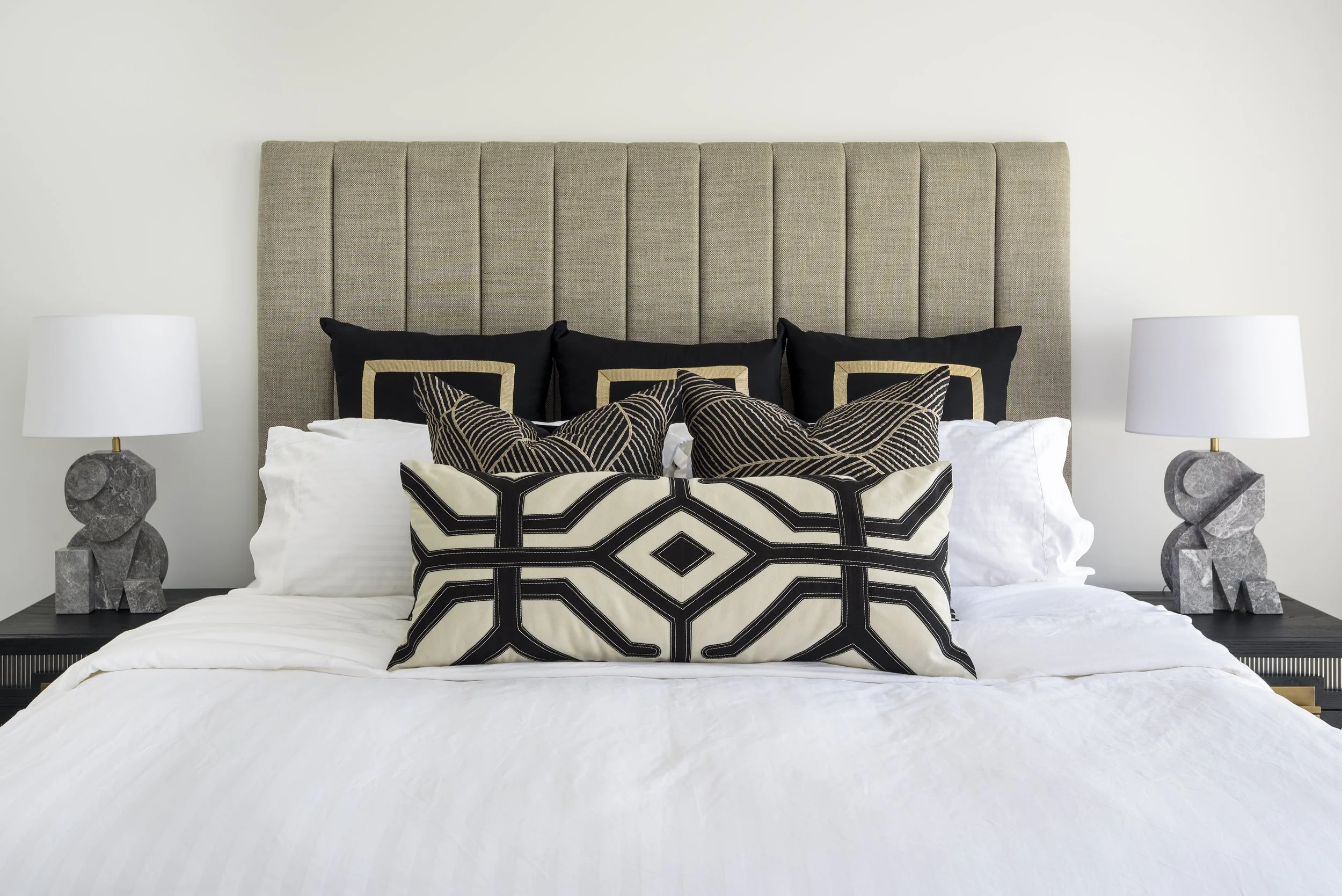 A neatly made bed with a tall beige upholstered headboard, white bedding, decorative black and gold pillows, and two small animal-shaped gray stone table lamps on black nightstands.