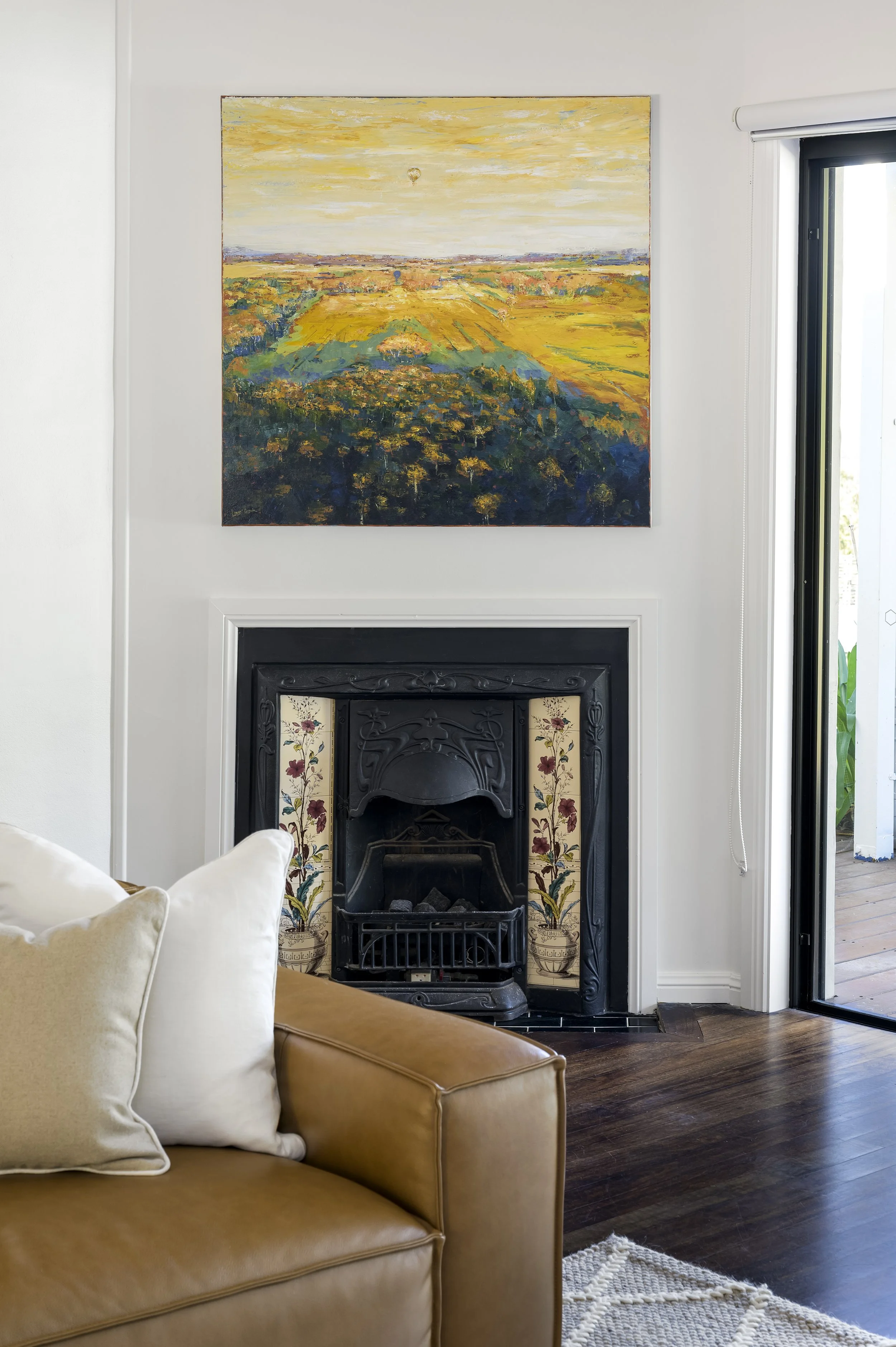 Interior of a living room featuring a black fireplace with floral tile detailing, a tan leather sofa with white pillows, a colorful landscape painting above the fireplace, and a sliding door leading outside.