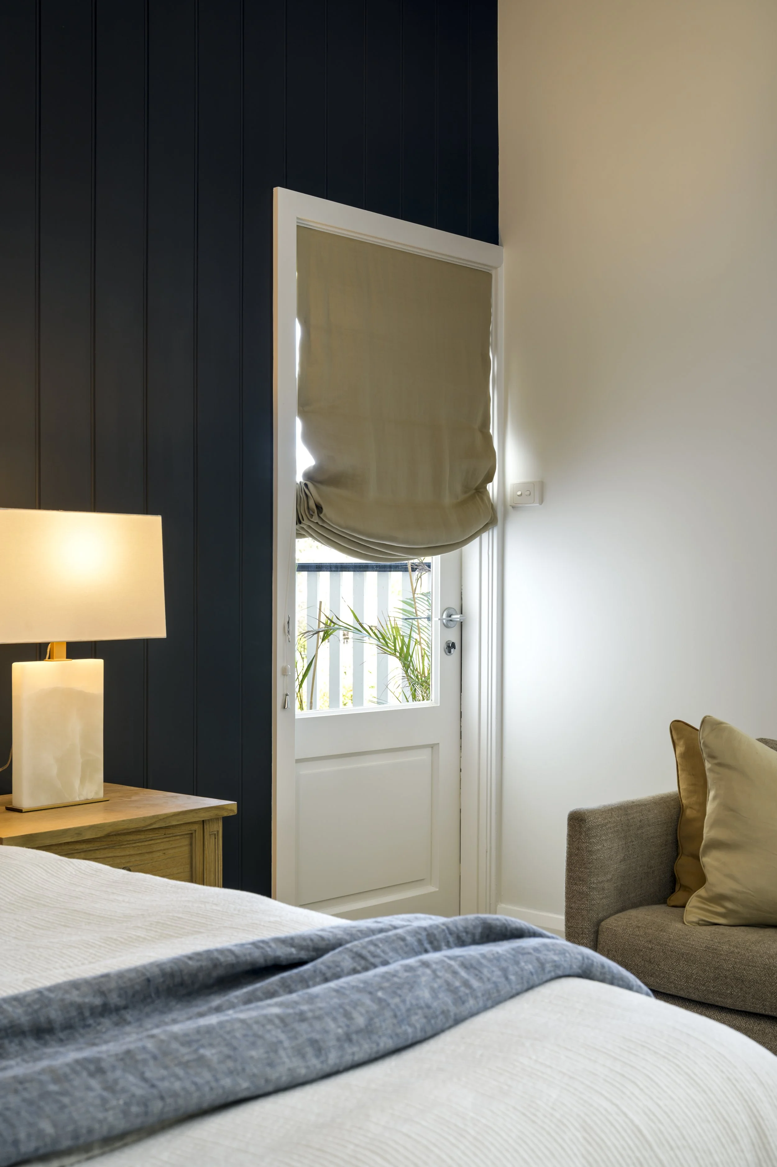 A cozy bedroom with a wooden nightstand, table lamp, bed with a gray blanket, a beige armchair with pillows, and a door with a beige Roman shade partially covering a window showing outdoor plants.
