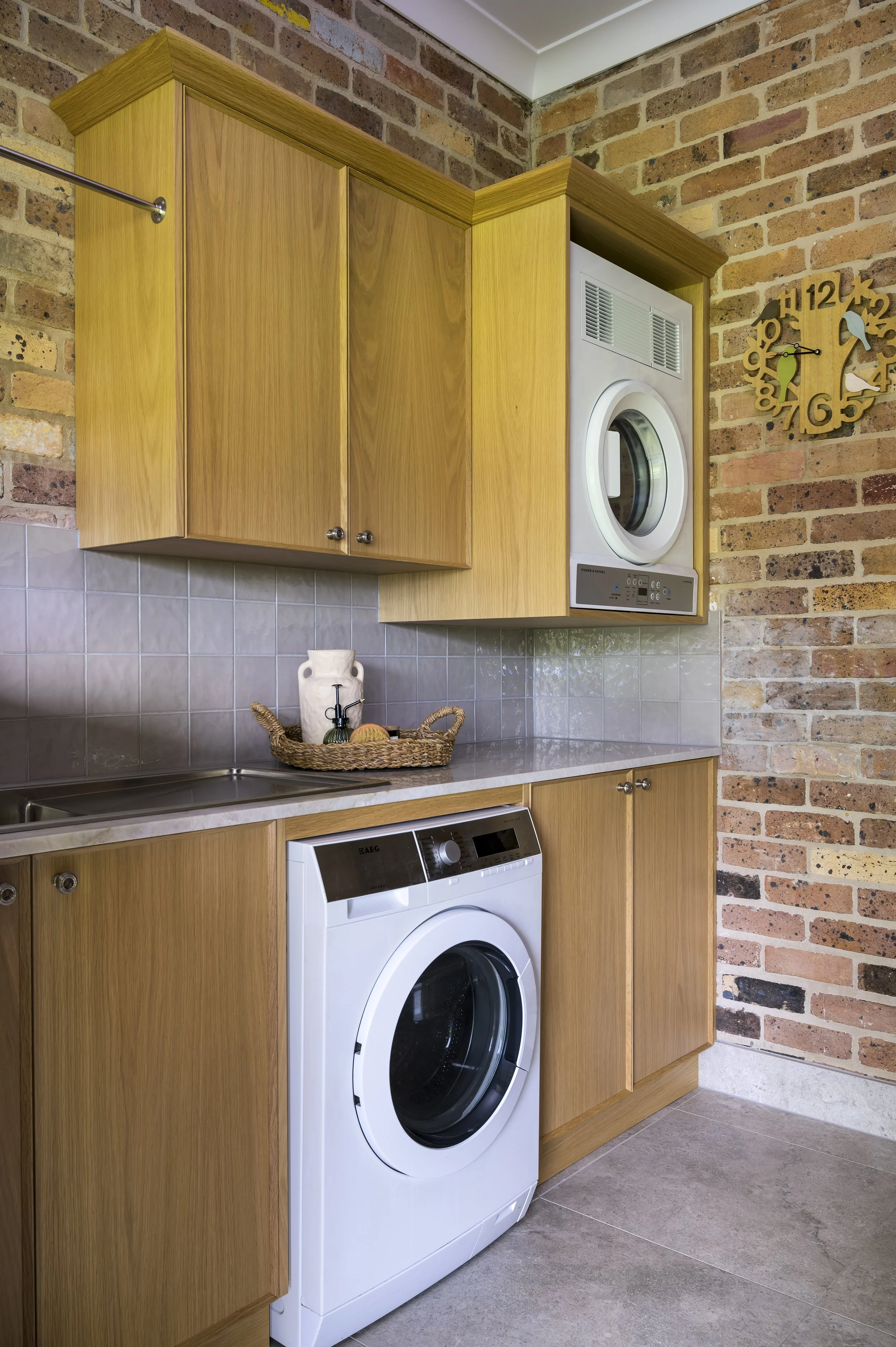 Laundry room with wooden cabinets, a washing machine, a laundry basket with soap and sponge, tiled backsplash, brick wall, and a decorative clock.