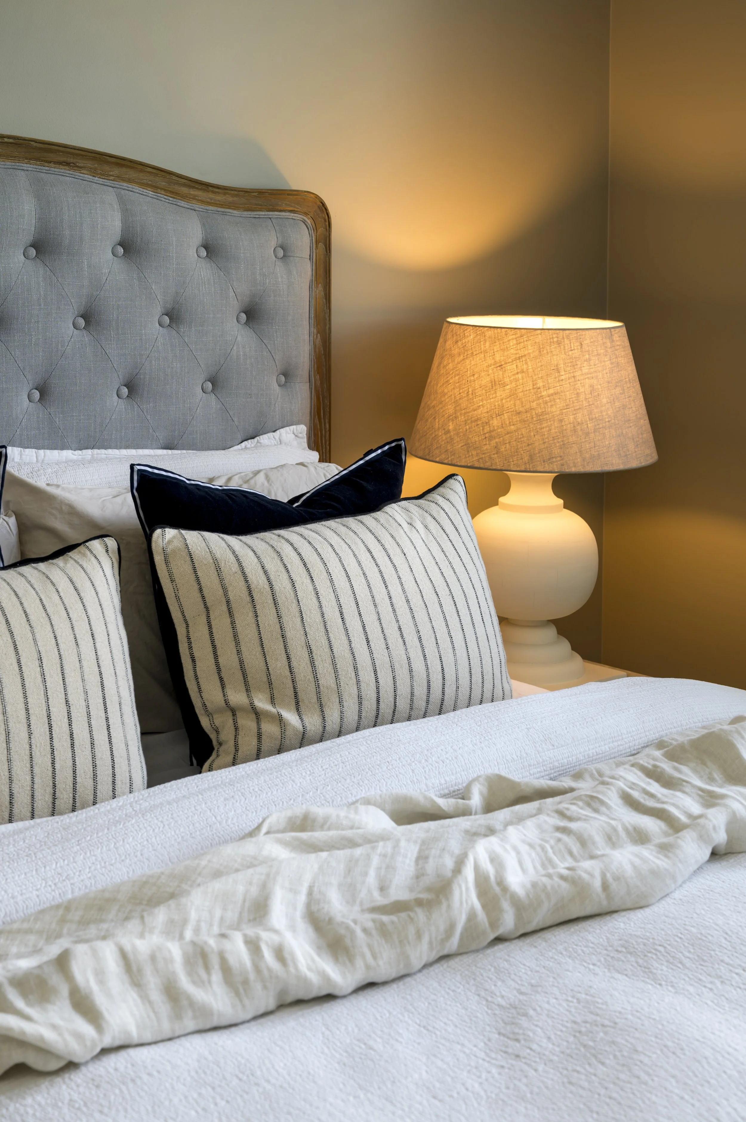 A cozy bedroom with a bed, striped pillows, a beige bedspread, a lampshade on a side table, and a softly lit wall in the background.