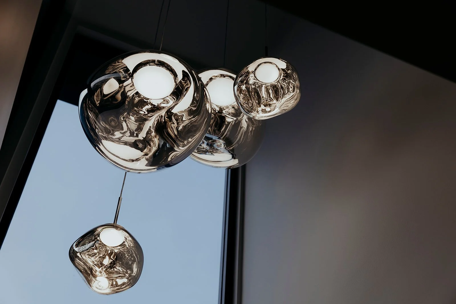 Modern metallic ceiling lights with reflective surfaces hanging near a large window, showing a clear sky.