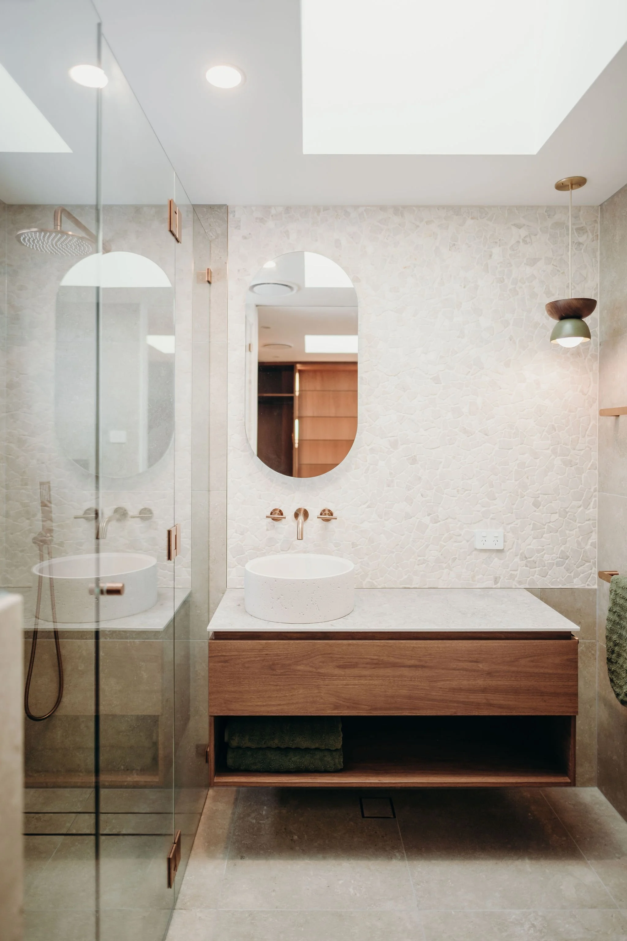 Modern bathroom with a wooden vanity, oval mirror, vessel sink, and a walk-in shower with glass door.