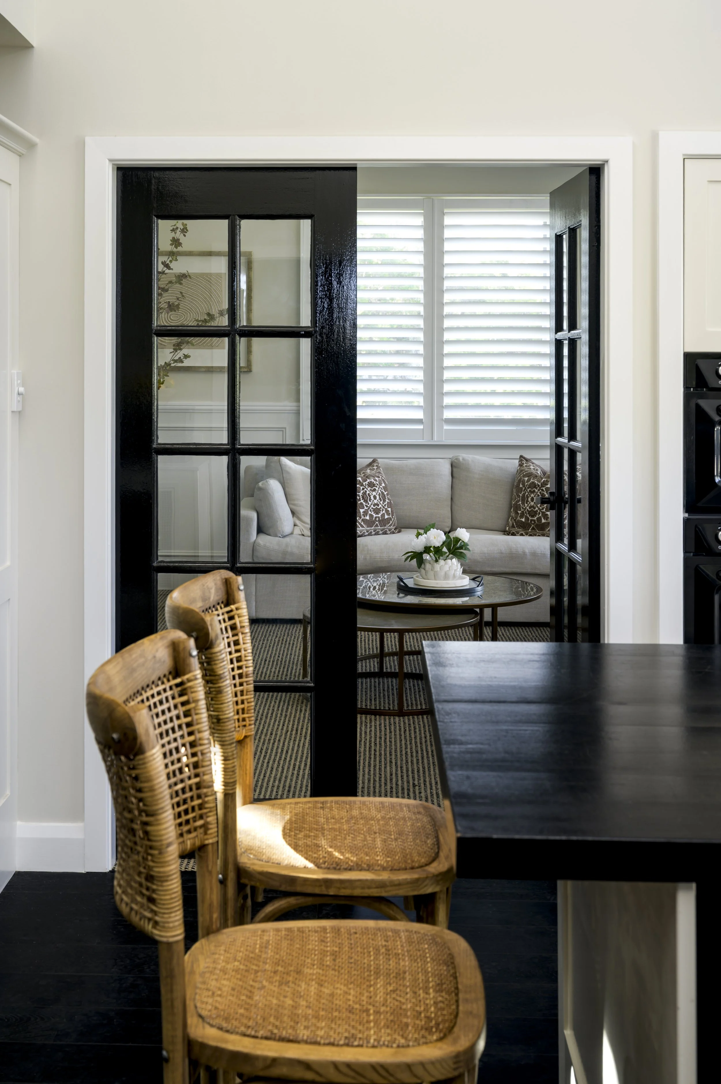 Interior view of a living room seen through black-framed glass doors, with a beige sofa, patterned pillows, a round coffee table with a white flower arrangement, and a window with plantation shutters.