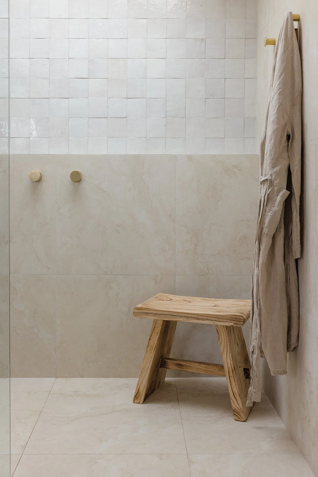 A minimalist bathroom shower area with beige tiles, a wooden stool, and a beige towel hanging on a golden hook.