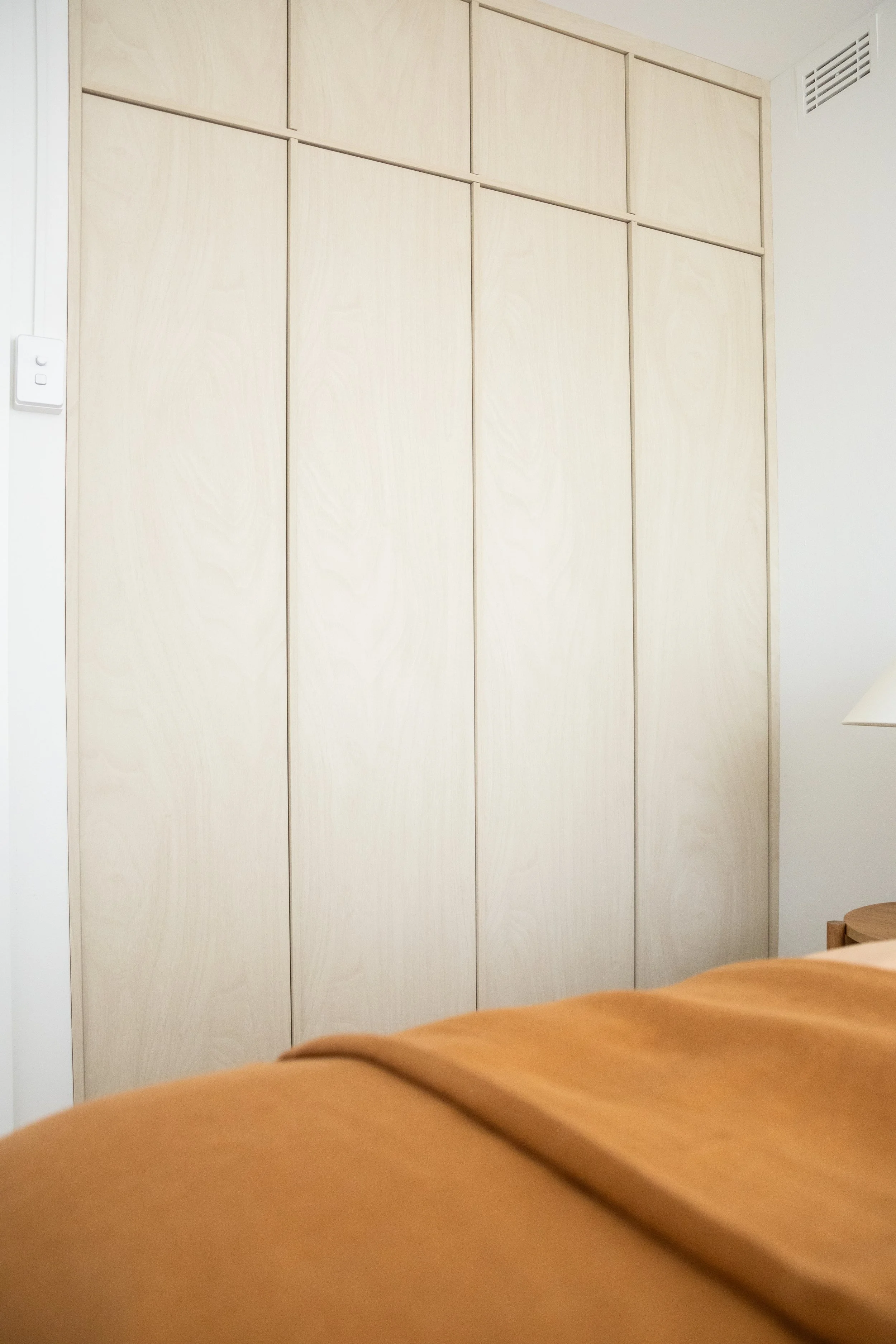 A wooden closet or cabinet with sliding doors in a light beige or natural wood finish, located in a bedroom with part of a bed and a nightstand visible.