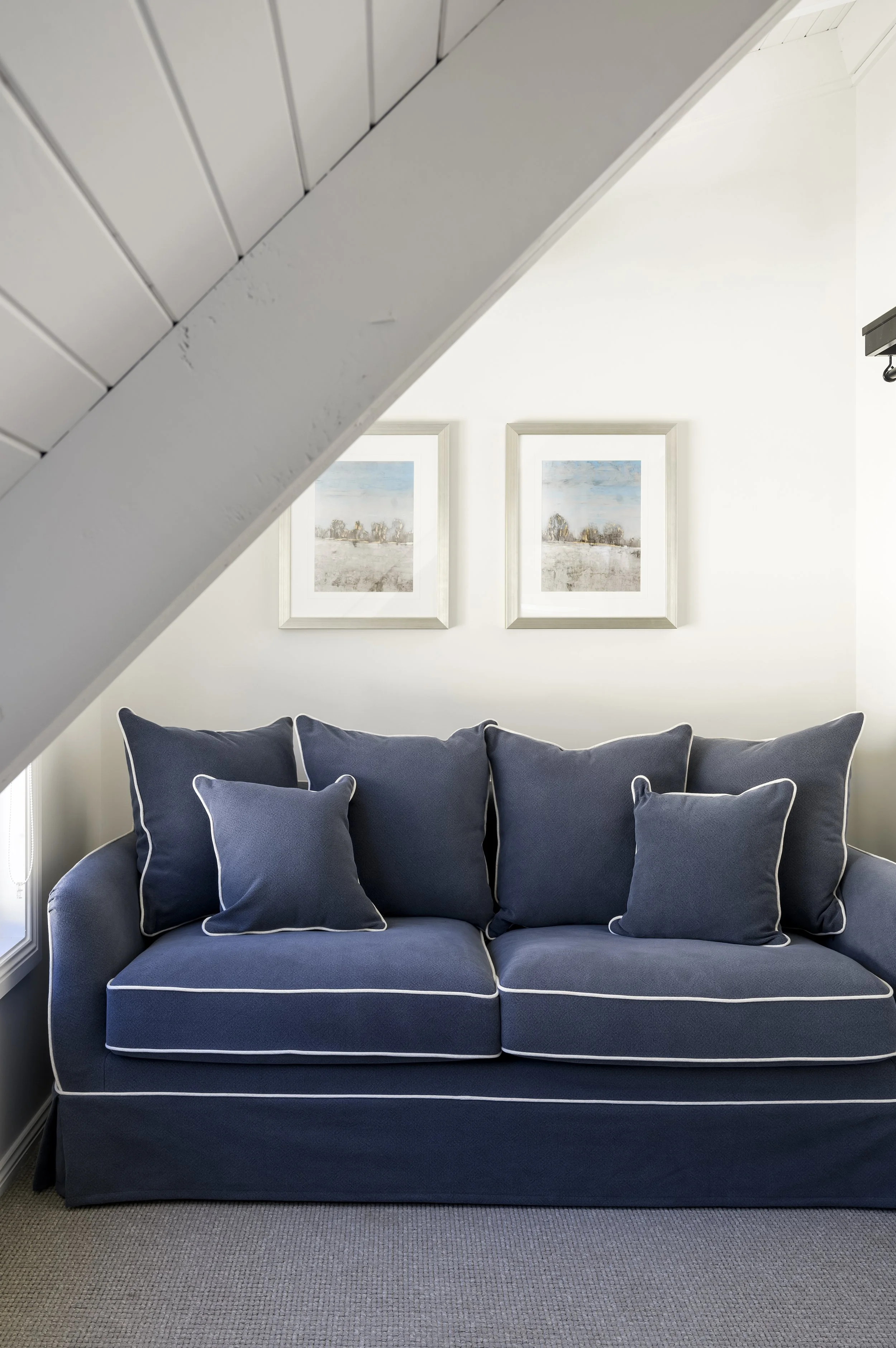 A blue sofa with multiple cushions in front of a white wall with two framed landscape paintings, partially obscured by a sloped ceiling.