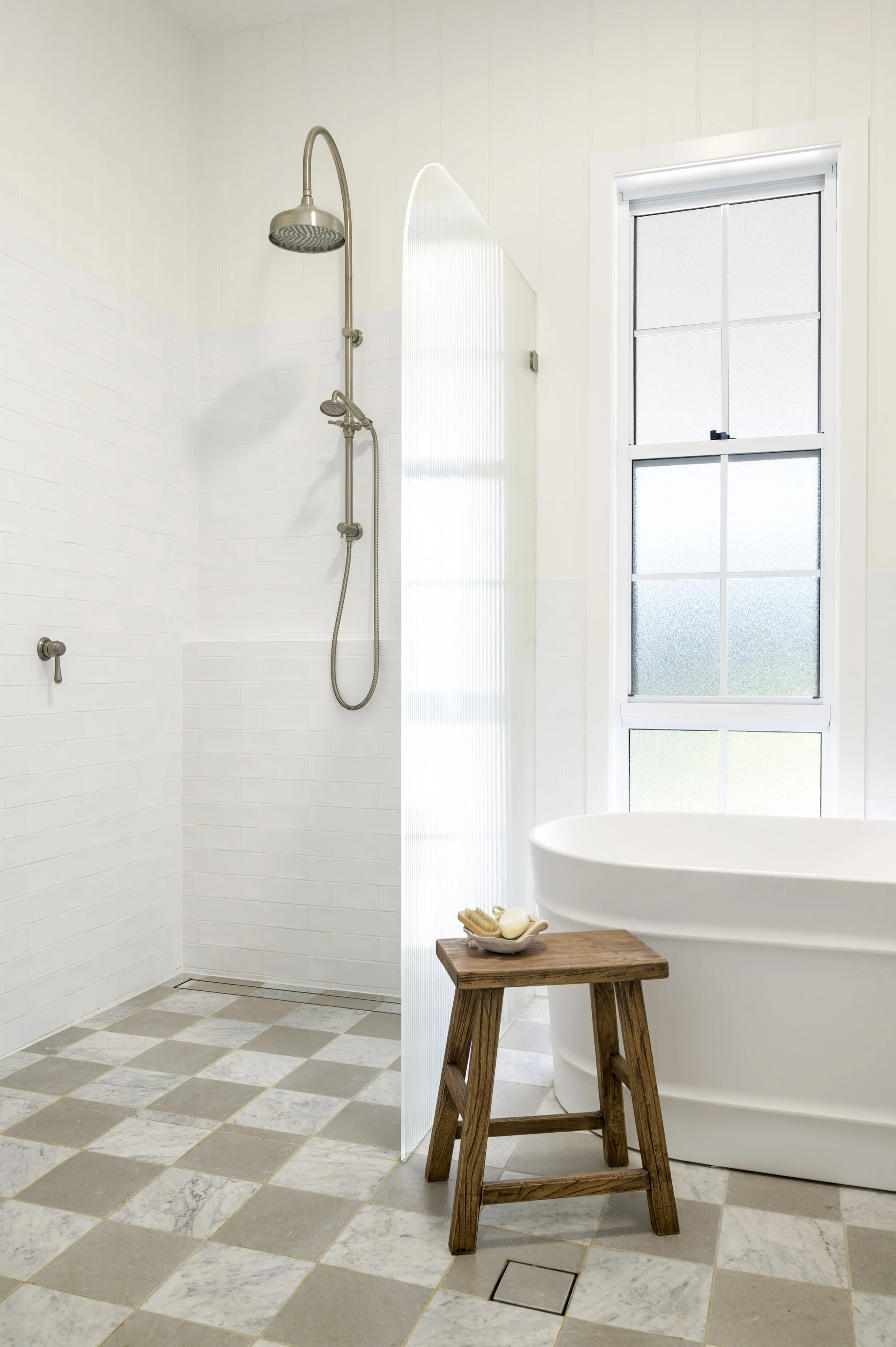 A modern bathroom with a walk-in shower, a white freestanding bathtub, a wooden stool with a soap dish, and a large window with frosted glass.