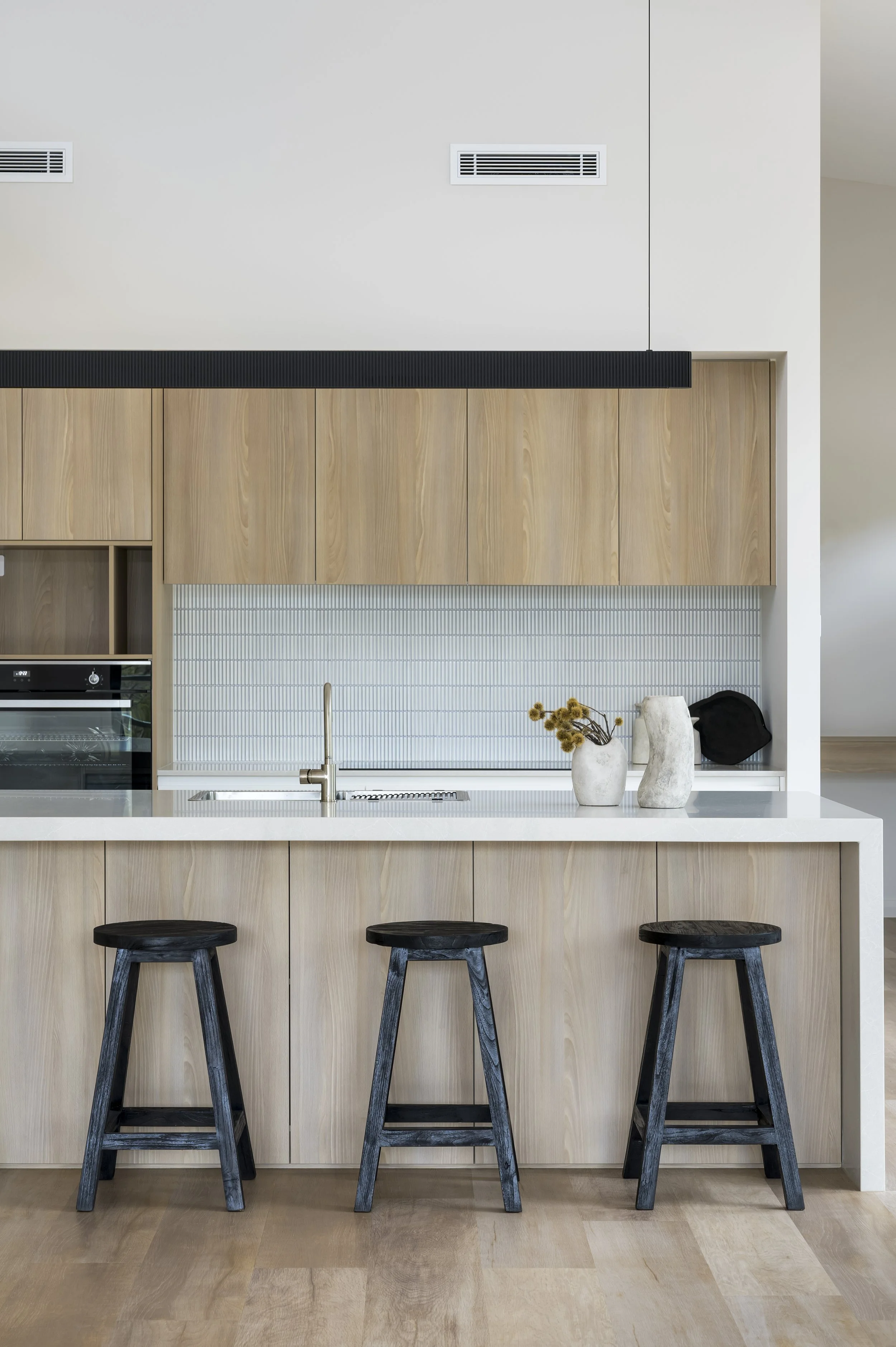 Modern kitchen with light wood cabinets, white countertop, black barstools, and decorative vases.