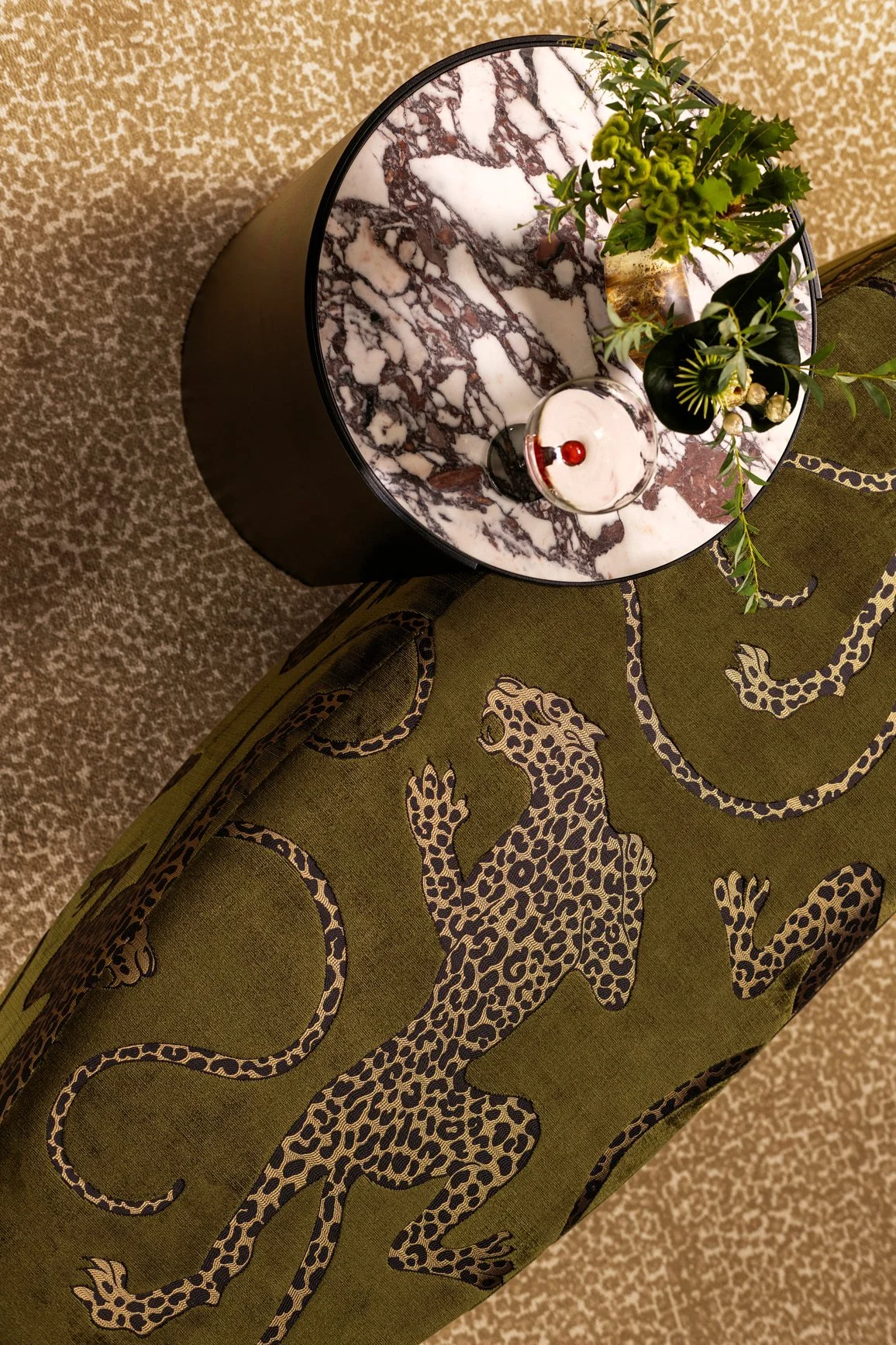 A round marble top black side table with a small plant in a glass jar and a glass of dark-colored beverage with red berries. The table is placed next to an upholstered chair with a green fabric featuring a pattern of leopards and their tails. The setting is on a beige speckled carpet.