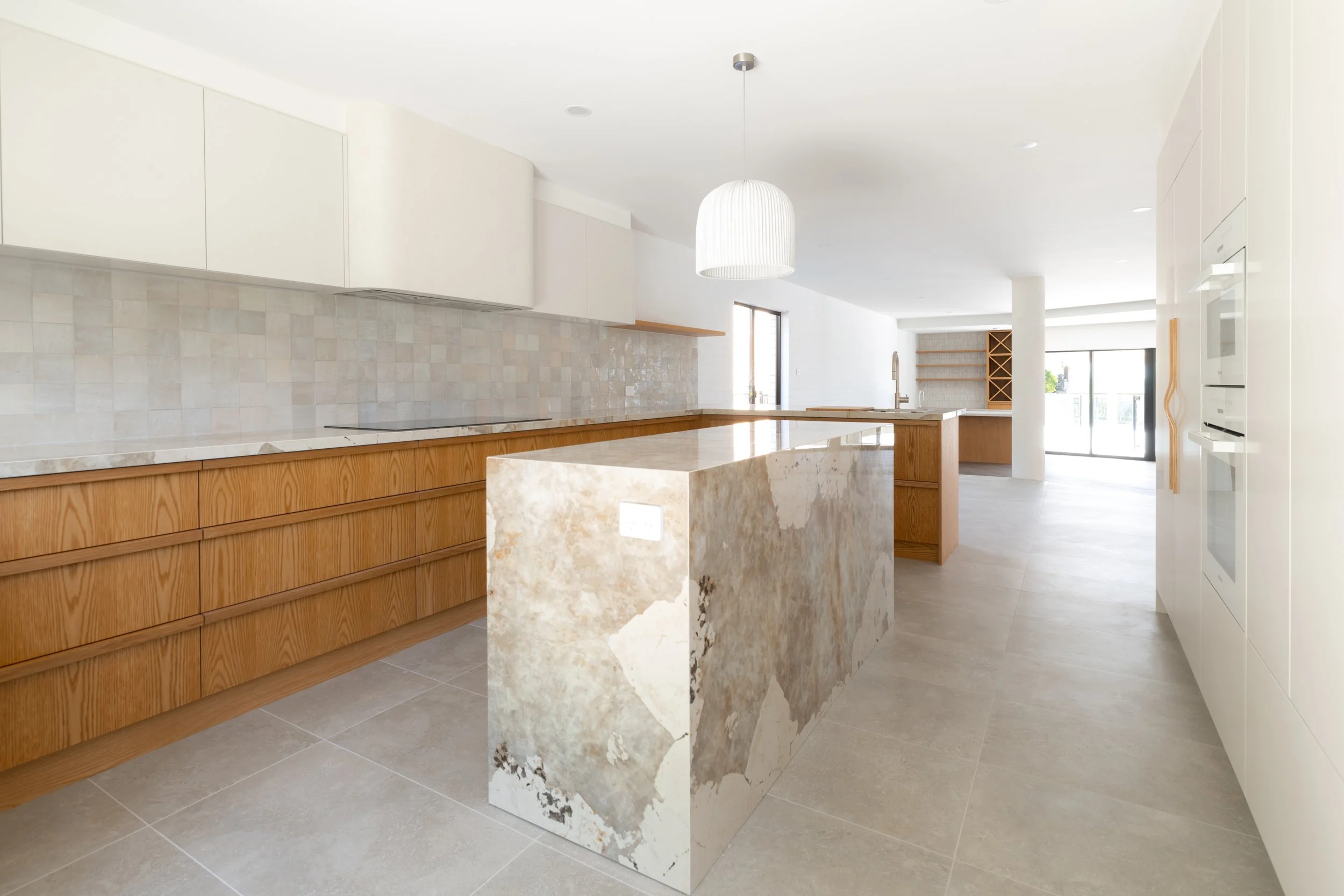 Modern kitchen with white and wooden cabinetry, a marble island, and tiled backsplash, with a large window and sliding door.