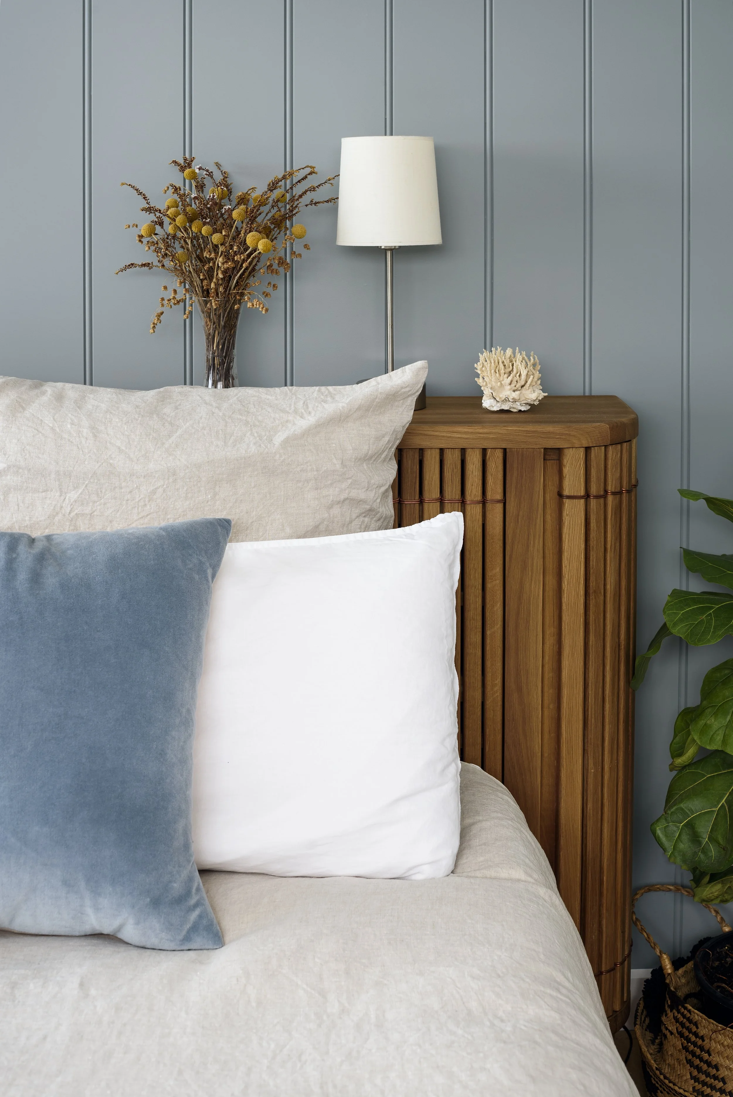 Close-up of a bedroom corner with beige, white, and blue pillows on a bed, a wooden nightstand with a white lamp, a vase with dried flowers, a decorative coral piece, and a potted plant against a blue paneled wall.