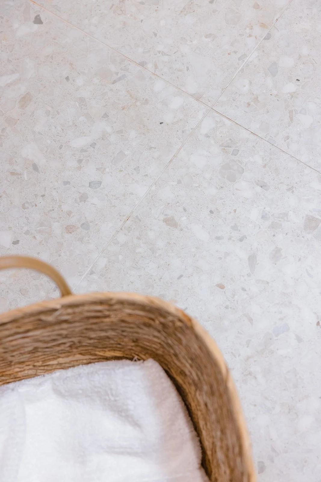 Part of a woven basket with a white towel inside on a light-colored tiled floor.