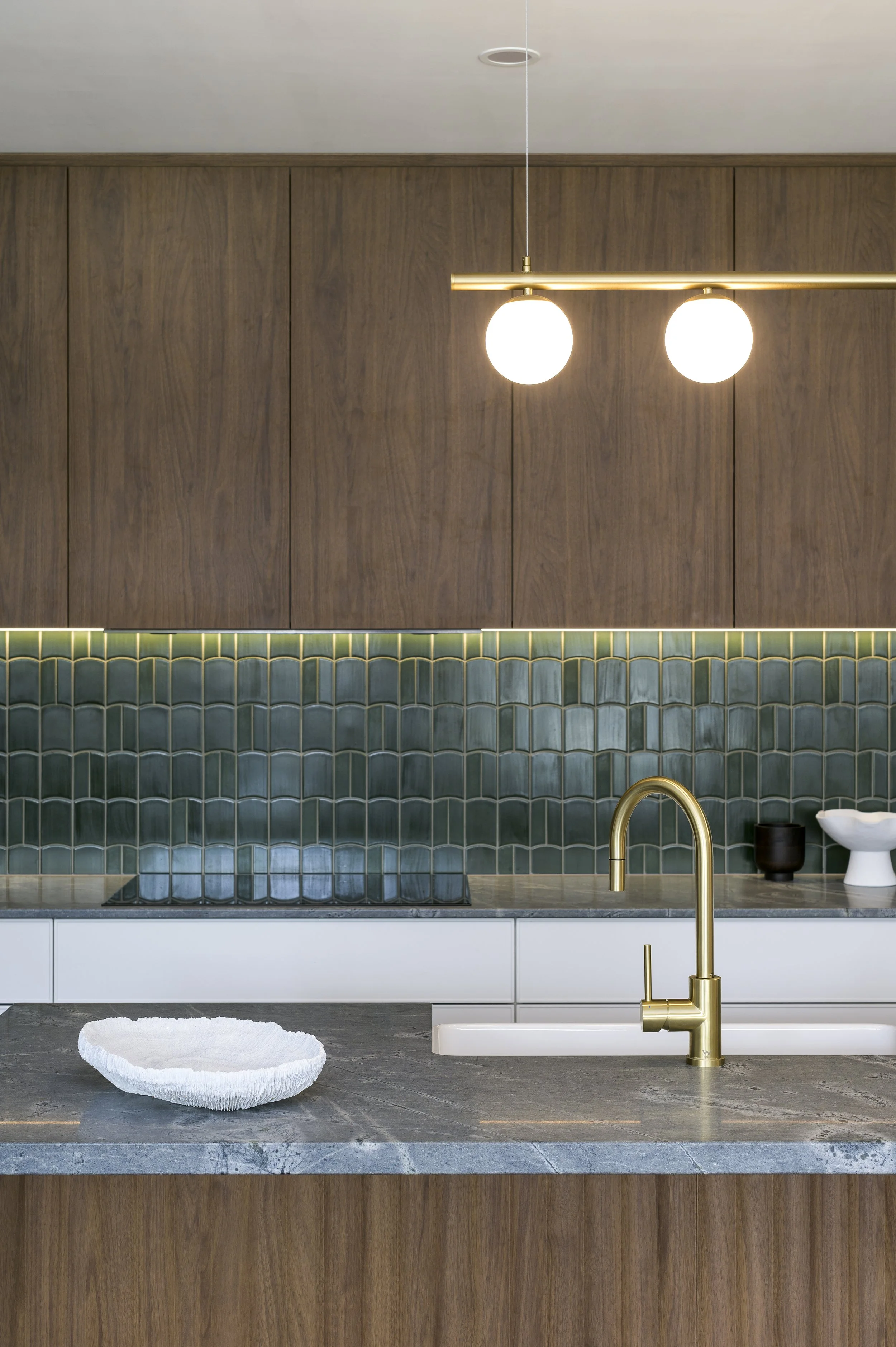 Modern kitchen with dark wood cabinets, green tiled backsplash, gold faucet, and a marble countertop with a white textured bowl.