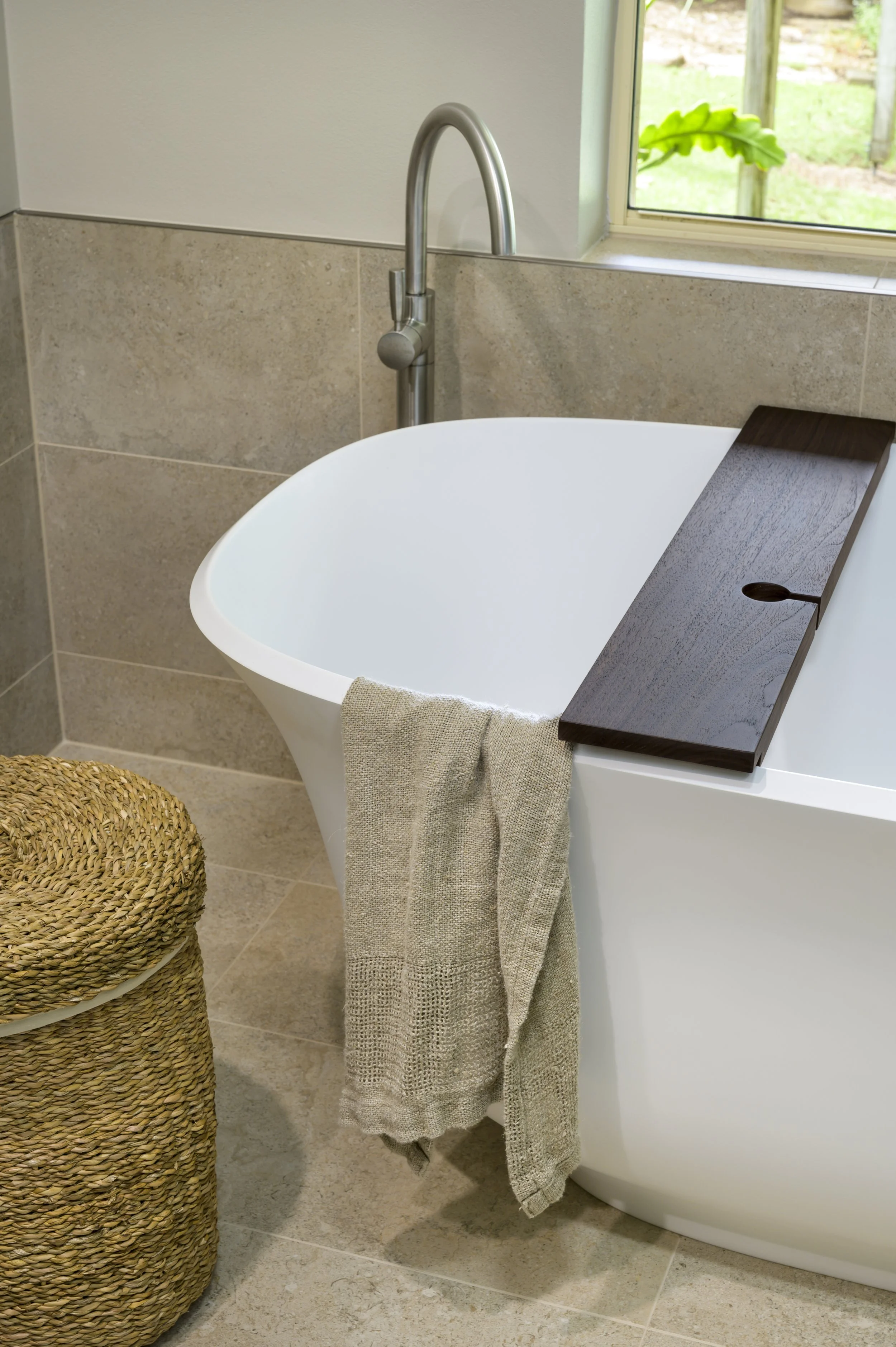 A white freestanding bathtub with a dark wooden bath tray, a beige towel hanging on the side, a woven laundry basket, a modern silver faucet, and a window with a view of green grass outside in a bathroom with beige tiled walls and floor.