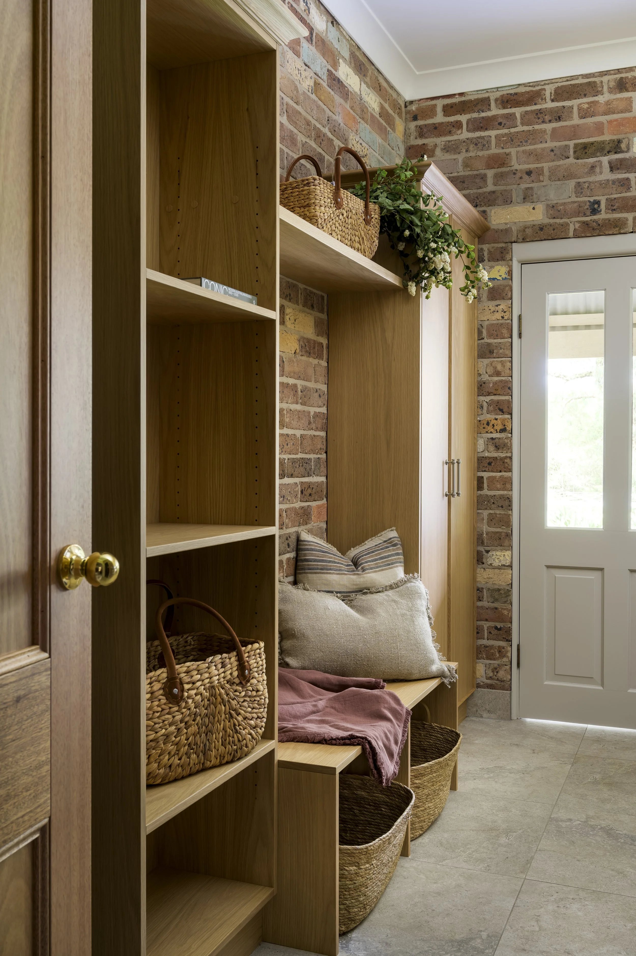 Entryway with wooden shelving, brick walls, baskets on the floor, pillows and a blanket on a bench, and a white door with glass panels.