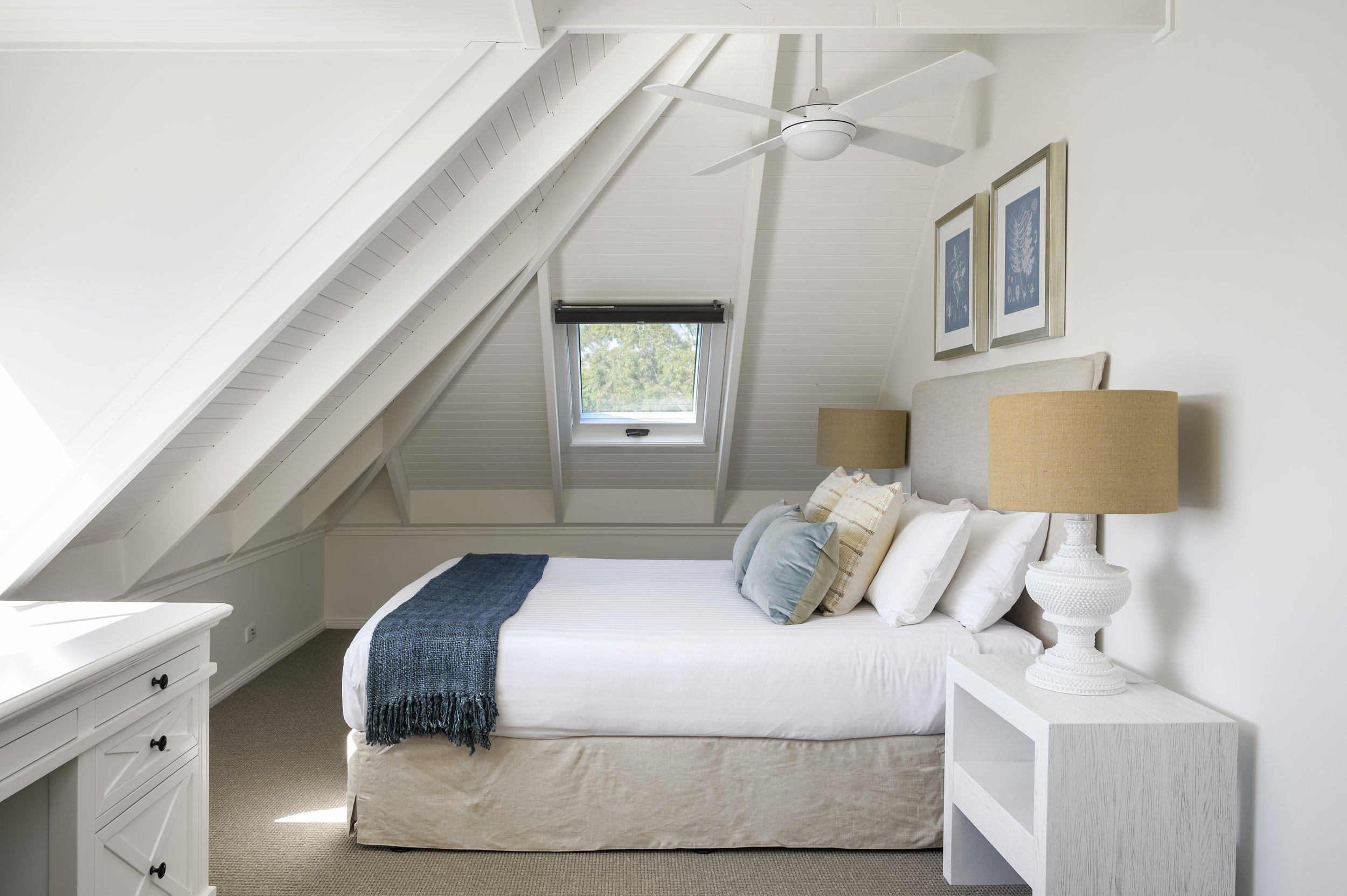 A cozy bedroom in an attic space with white walls and sloped ceiling. It features a bed with white bedding, beige skirt, and decorative pillows. There is a white nightstand with a textured white table lamp, a small window with a black blind, and framed artwork on the wall above the bed. A ceiling fan with white blades is mounted on the ceiling.