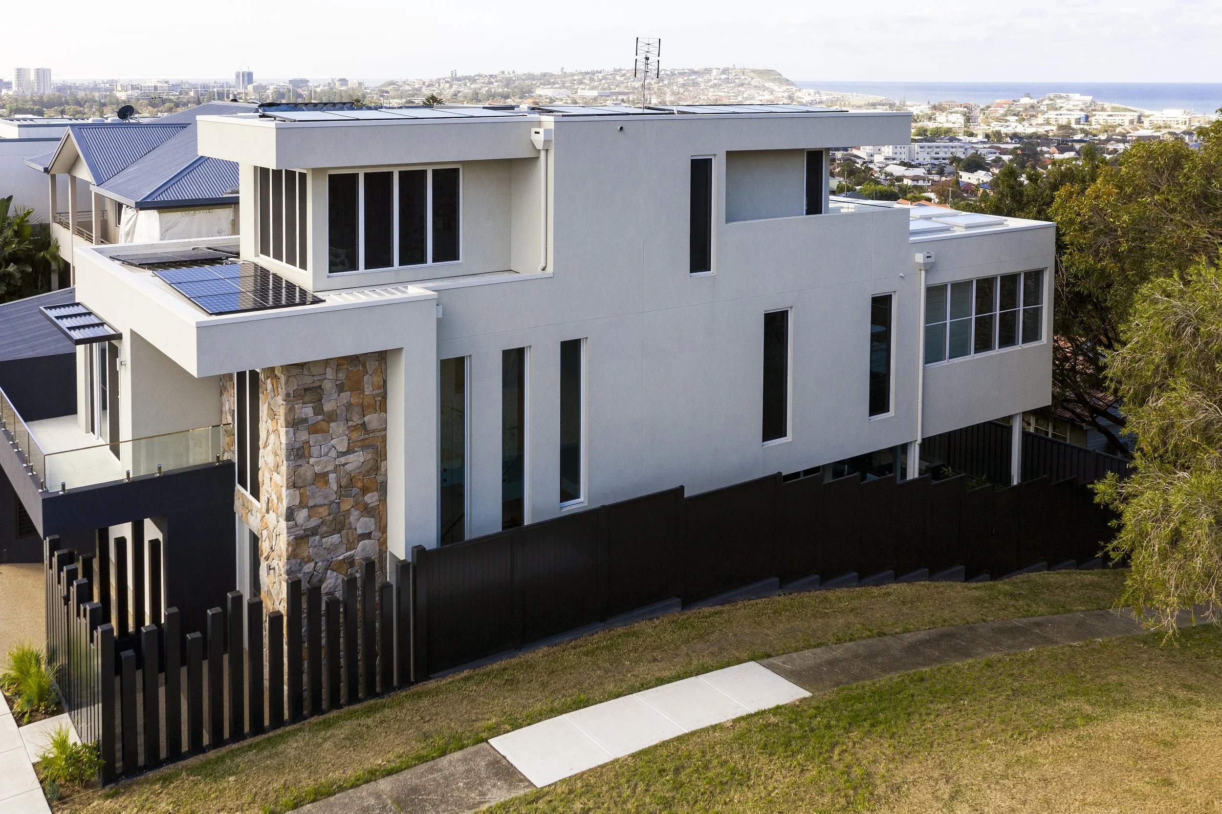 Modern multi-story house with white exterior, large windows, stone accent wall, solar panels on roof, and a black fence in a hilly area.