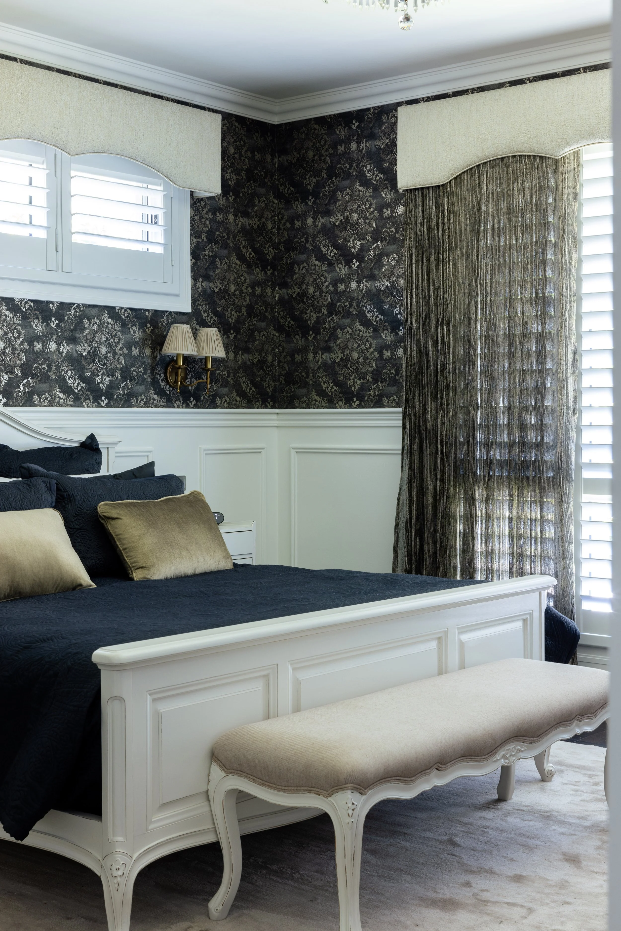A bedroom with large windows, dark floral wallpaper on the upper walls, white wainscoting, a white bed with black bedding and gold pillows, a beige bench at the foot of the bed, and curtains covering the windows.