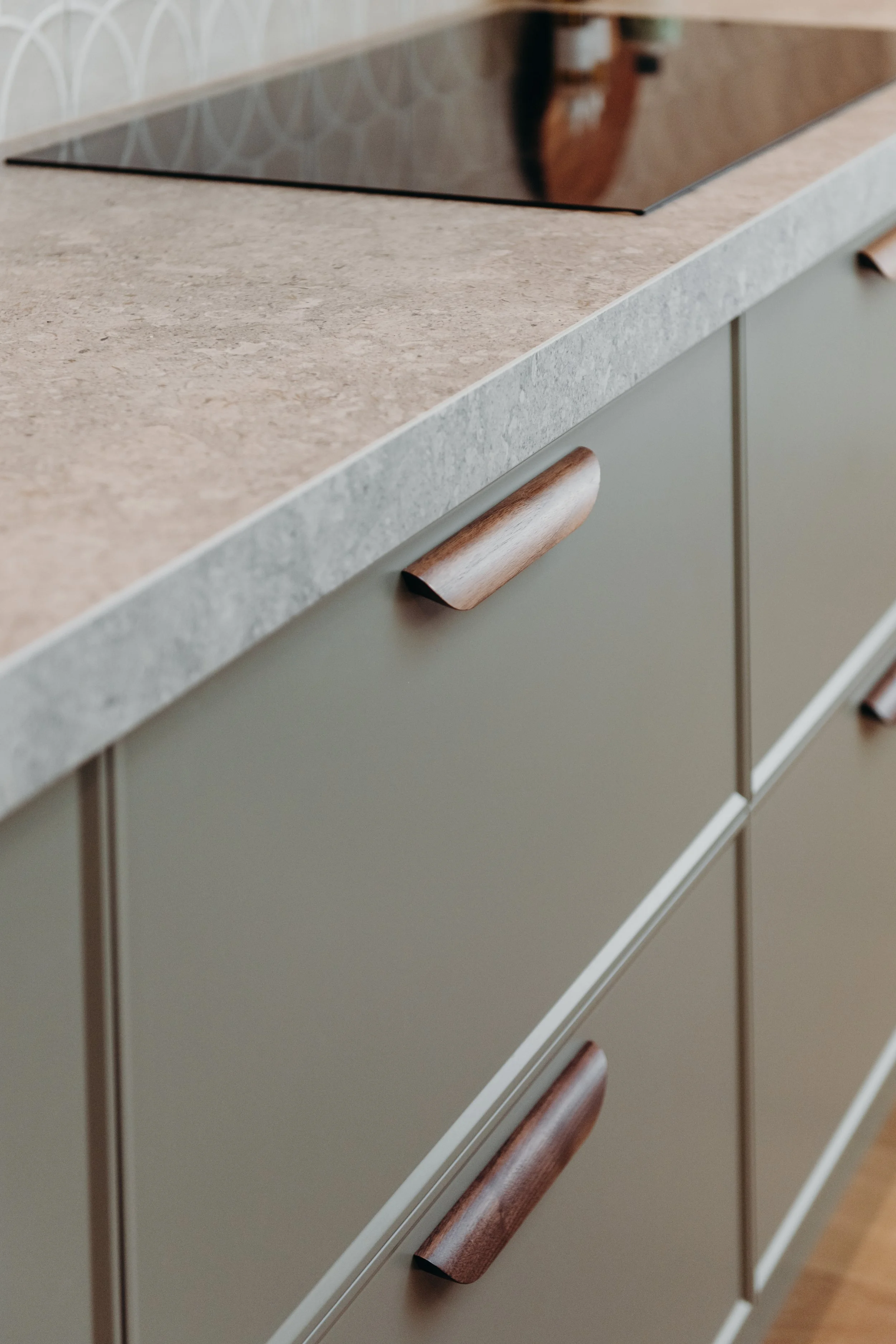 Close-up of kitchen cabinets with beige doors and horizontal wooden handles, and a gray marble countertop with a black ceramic cooktop.