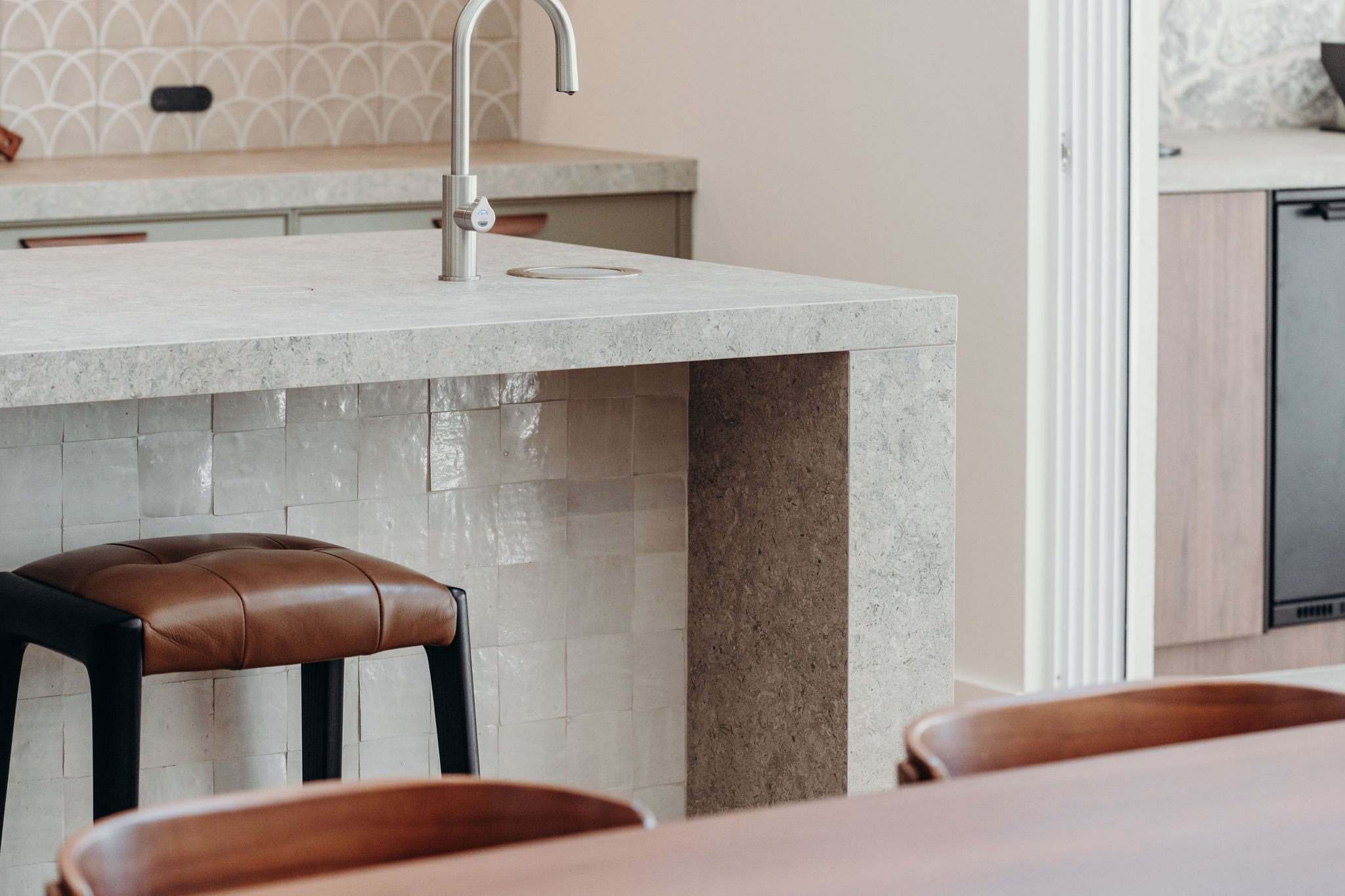 Modern kitchen with a light-colored stone countertop, a stainless steel faucet, and brown and black chairs around a wooden table.