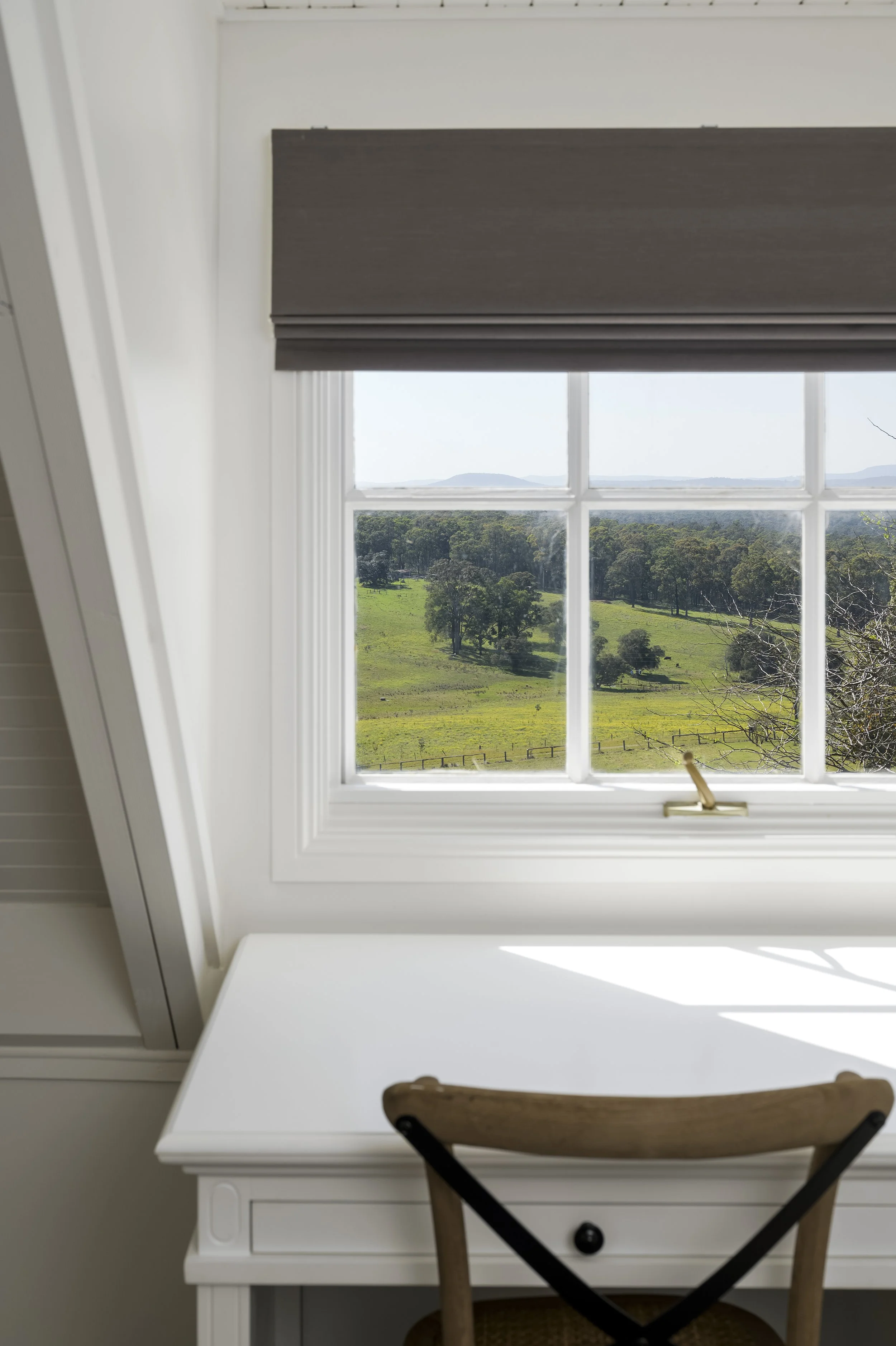 A white desk and wooden chair facing a large window showing a green hilly landscape with trees and distant mountains.