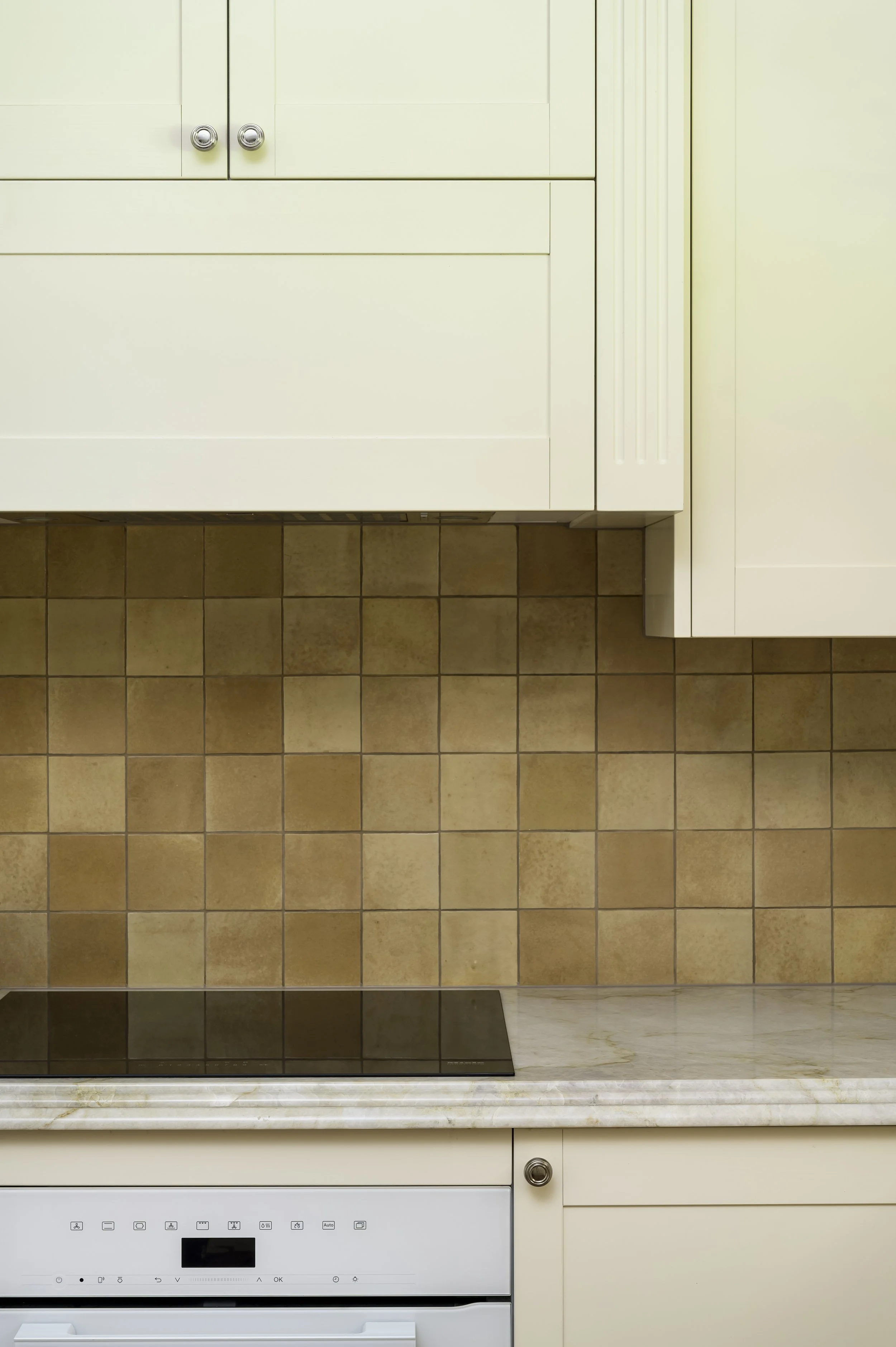 Close-up of a classic kitchen with white cabinets, a marble countertop, brown tiled backsplash, and a black electric cooktop.