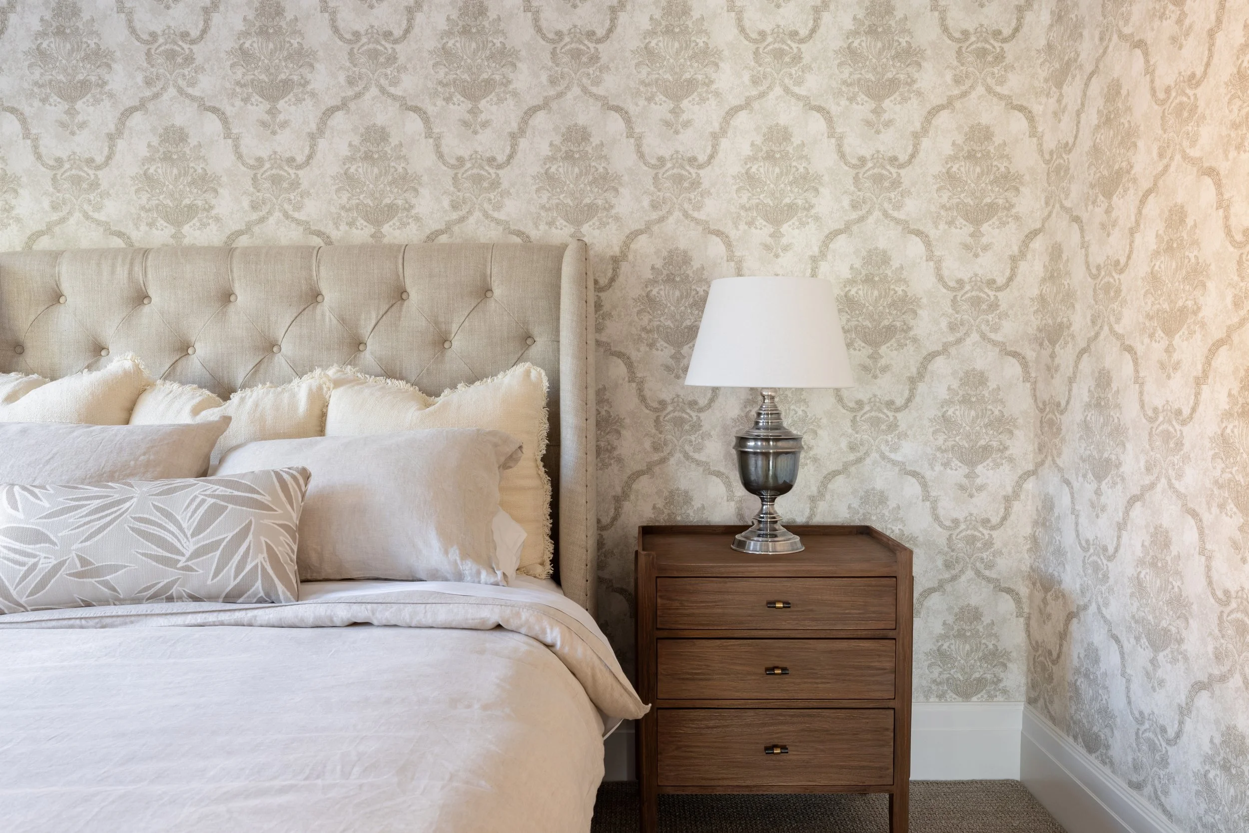 A neatly made bed with beige and cream-colored pillows and comforter, adjacent to a wooden nightstand with three drawers and a silver and white table lamp, against a wall with cream and beige damask wallpaper.