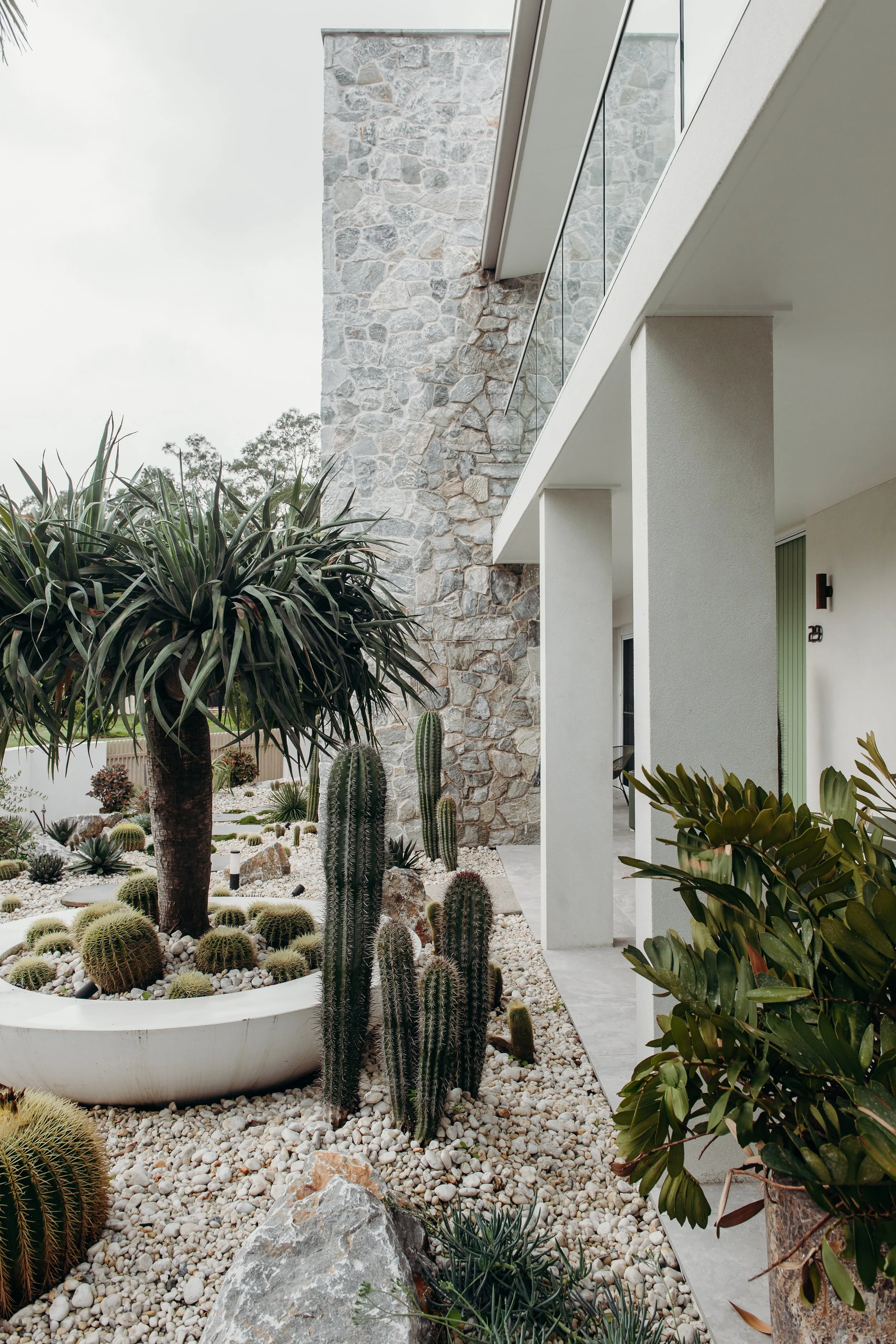 Modern house exterior with stone wall, white pillars, and enclosed patio featuring desert plants including cacti and succulents in white planters and gravel.