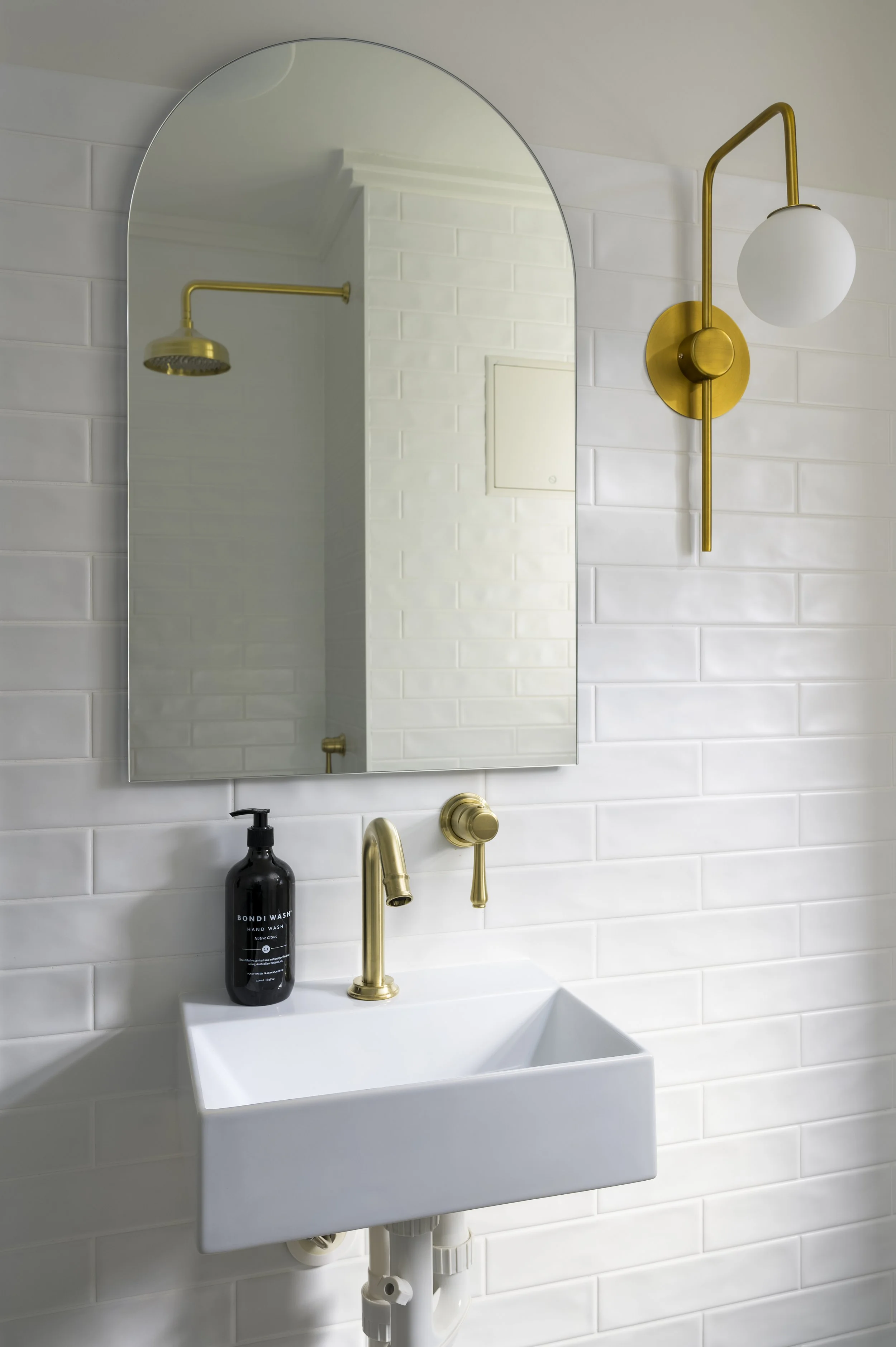 A modern white bathroom sink with gold fixtures, a black hand soap dispenser, a mirror, and a brass wall-mounted light