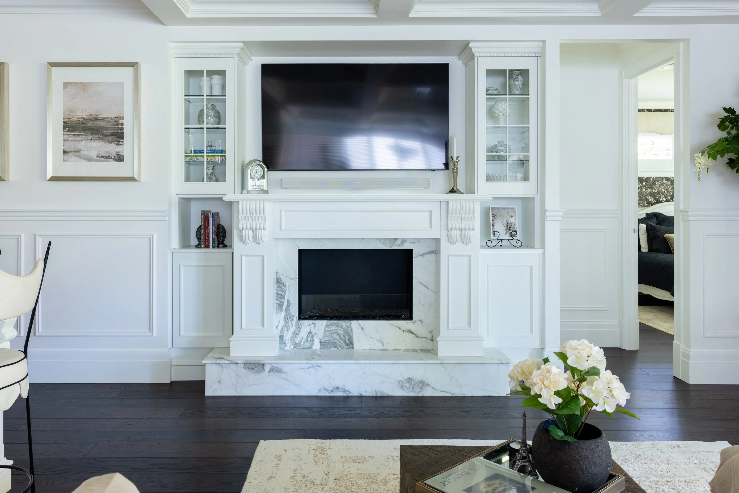 Modern living room with a white fireplace, mounted TV, and built-in shelves, decorated with vases, books, and framed artwork. A coffee table with a potted white flower and decorative items, and a doorway leading to a bedroom with dark bedding.