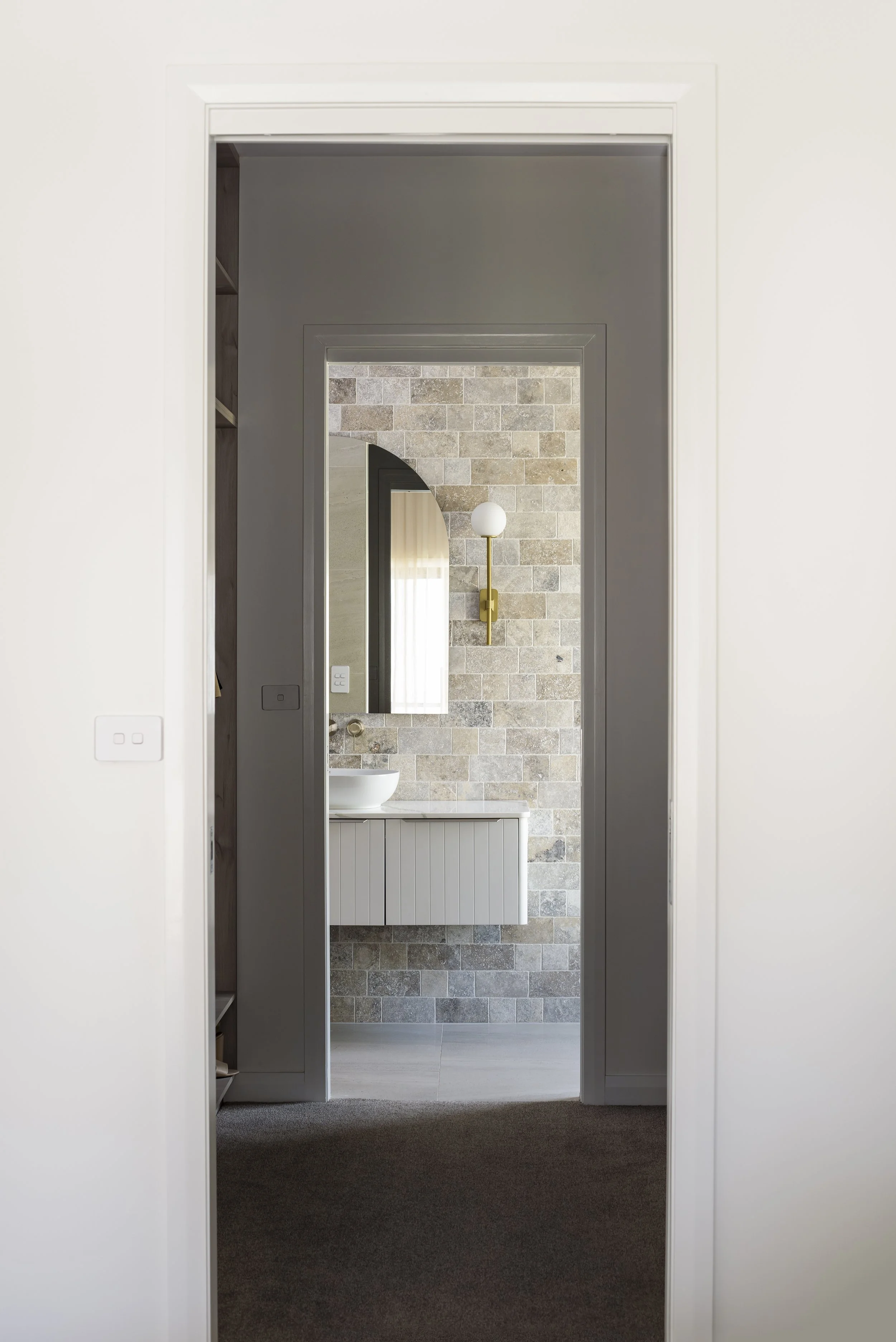 View through doorway into bathroom with stone-tile wall, mirror, white sink, and modern brass wall sconce.