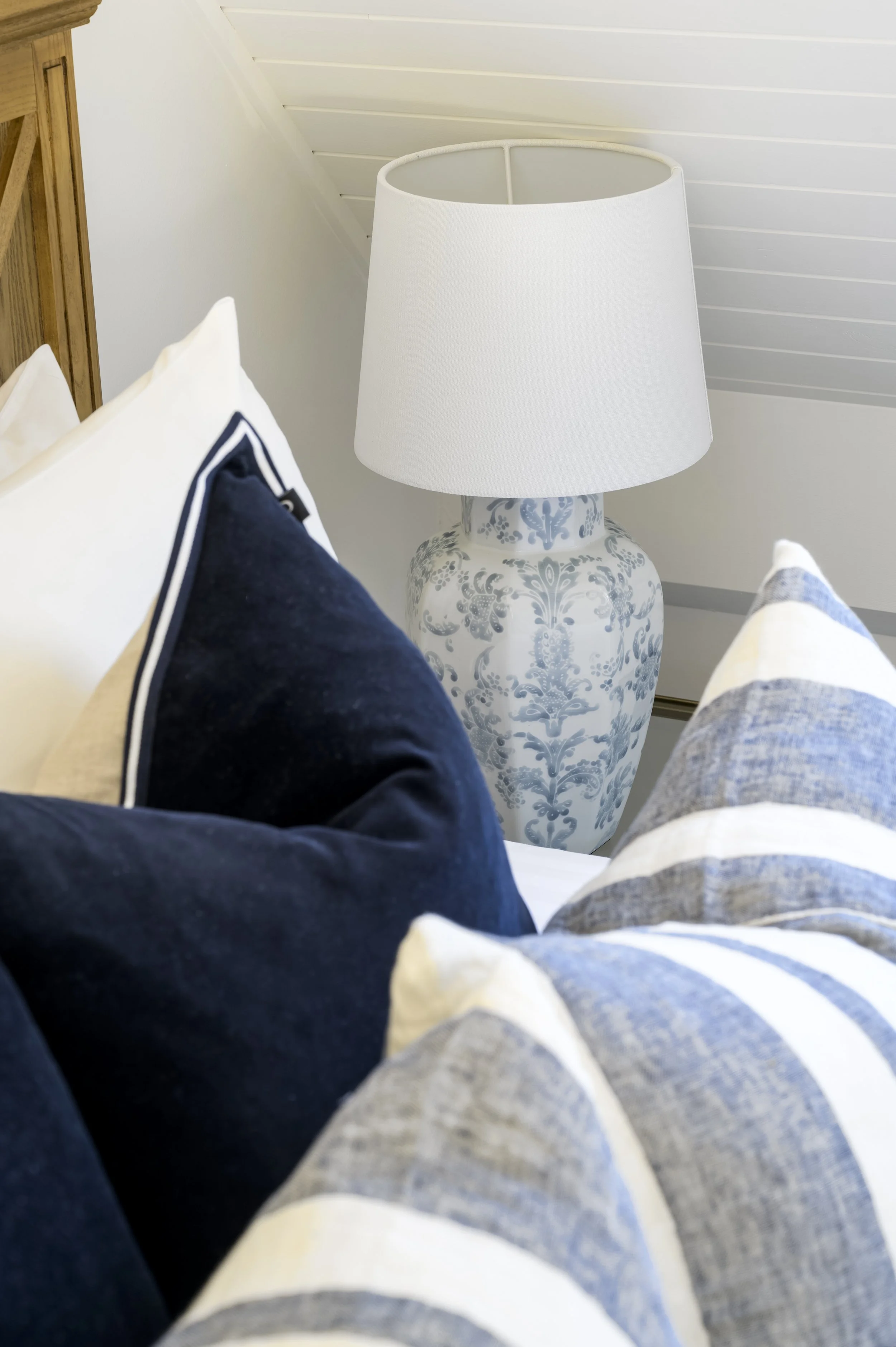 Clothes and pillows on a bed with a decorative vase and lampshade in the background.